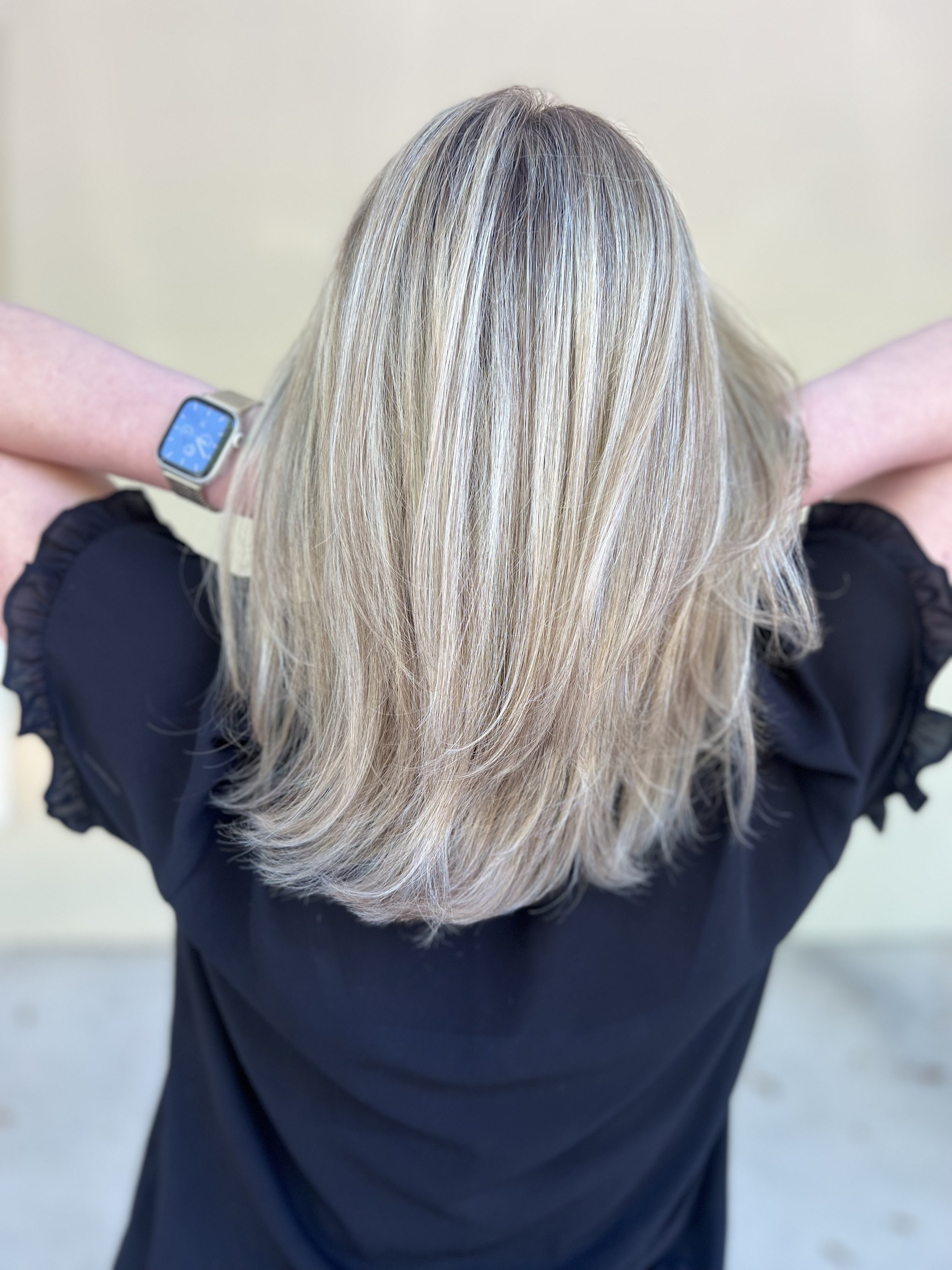 Back view of a woman with shoulder-length blonde hair wearing a black top and a silver smartwatch, standing outdoors against a light-colored wall.