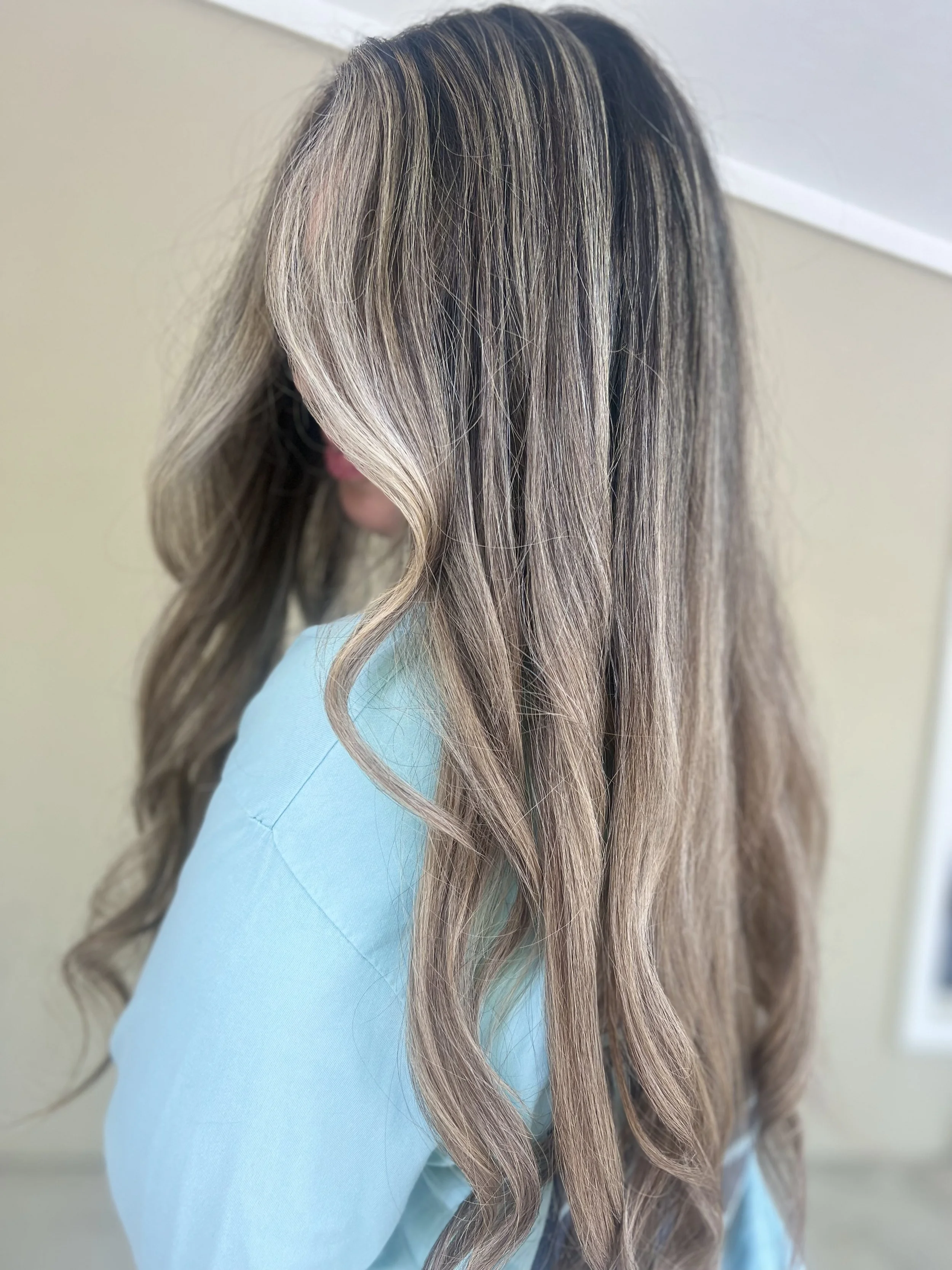 Woman with long, wavy, blonde hair with darker roots, wearing a light blue top.