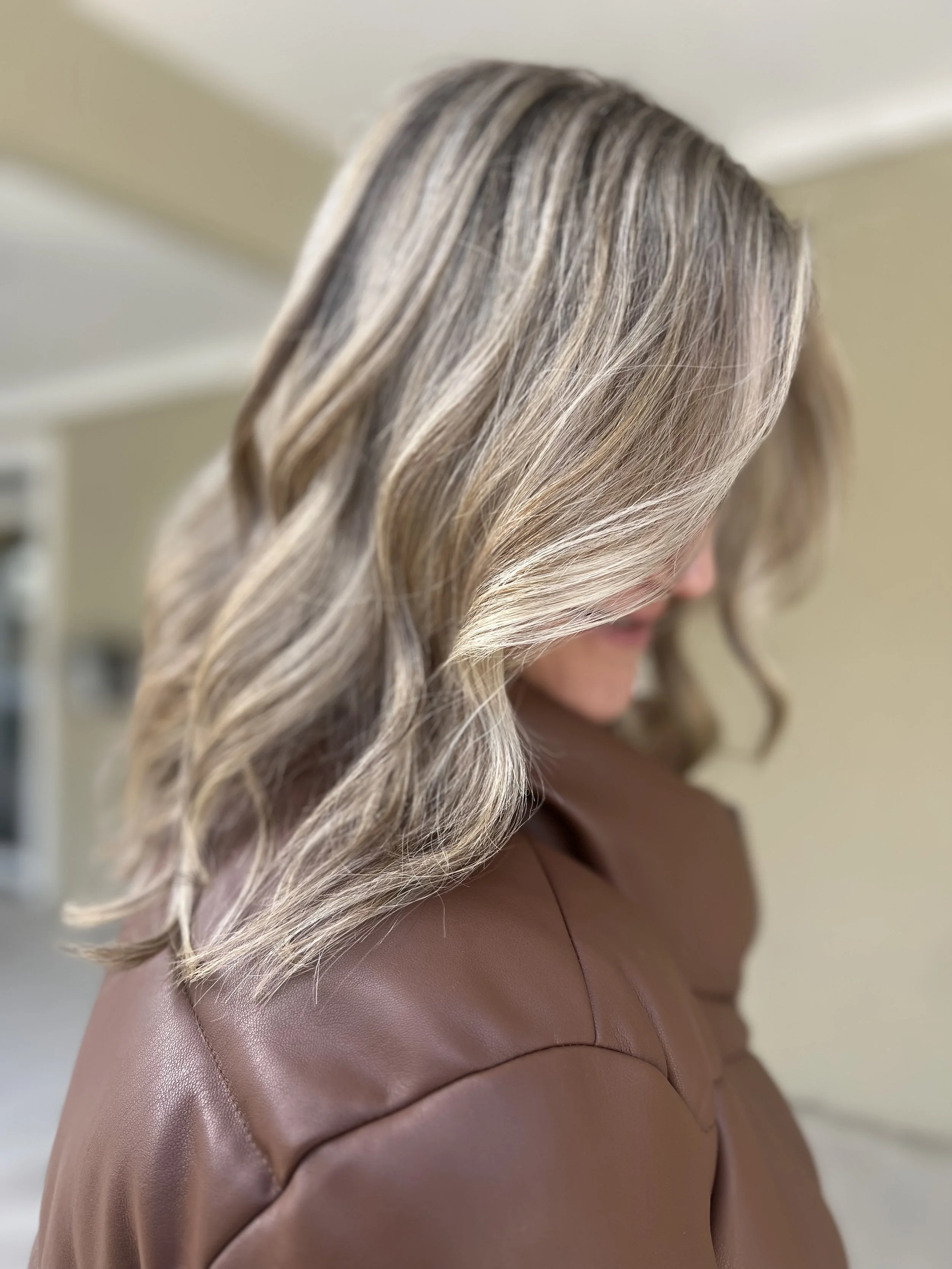 Close-up of a woman with blonde, wavy hair wearing a brown leather jacket, looking down, in an indoor setting.