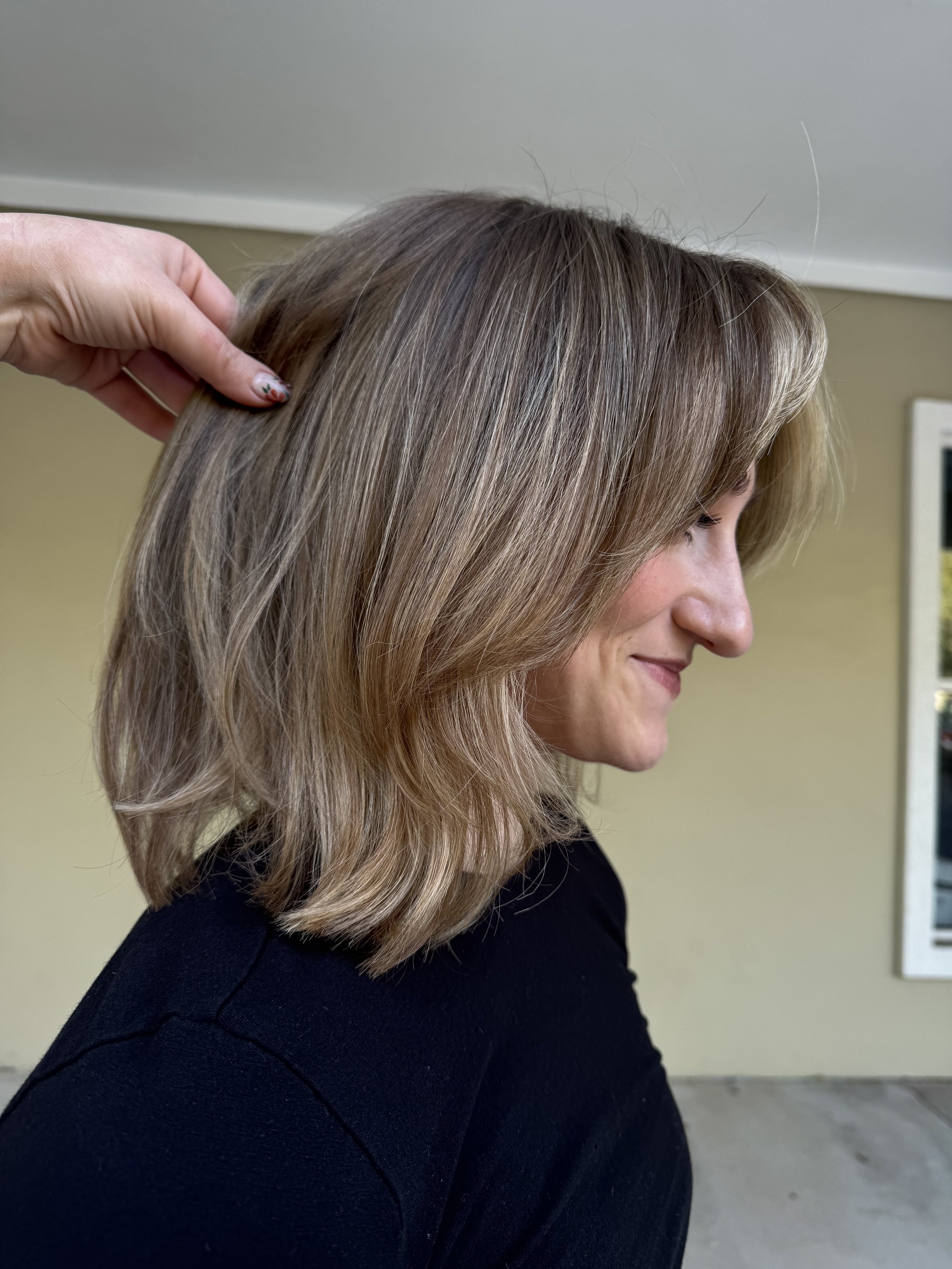 A woman with shoulder-length light brown hair, styled with soft waves, viewed from the side, with a hand touching her hair from the left. She is smiling slightly and wearing a black top, standing in front of a beige wall with a framed picture.