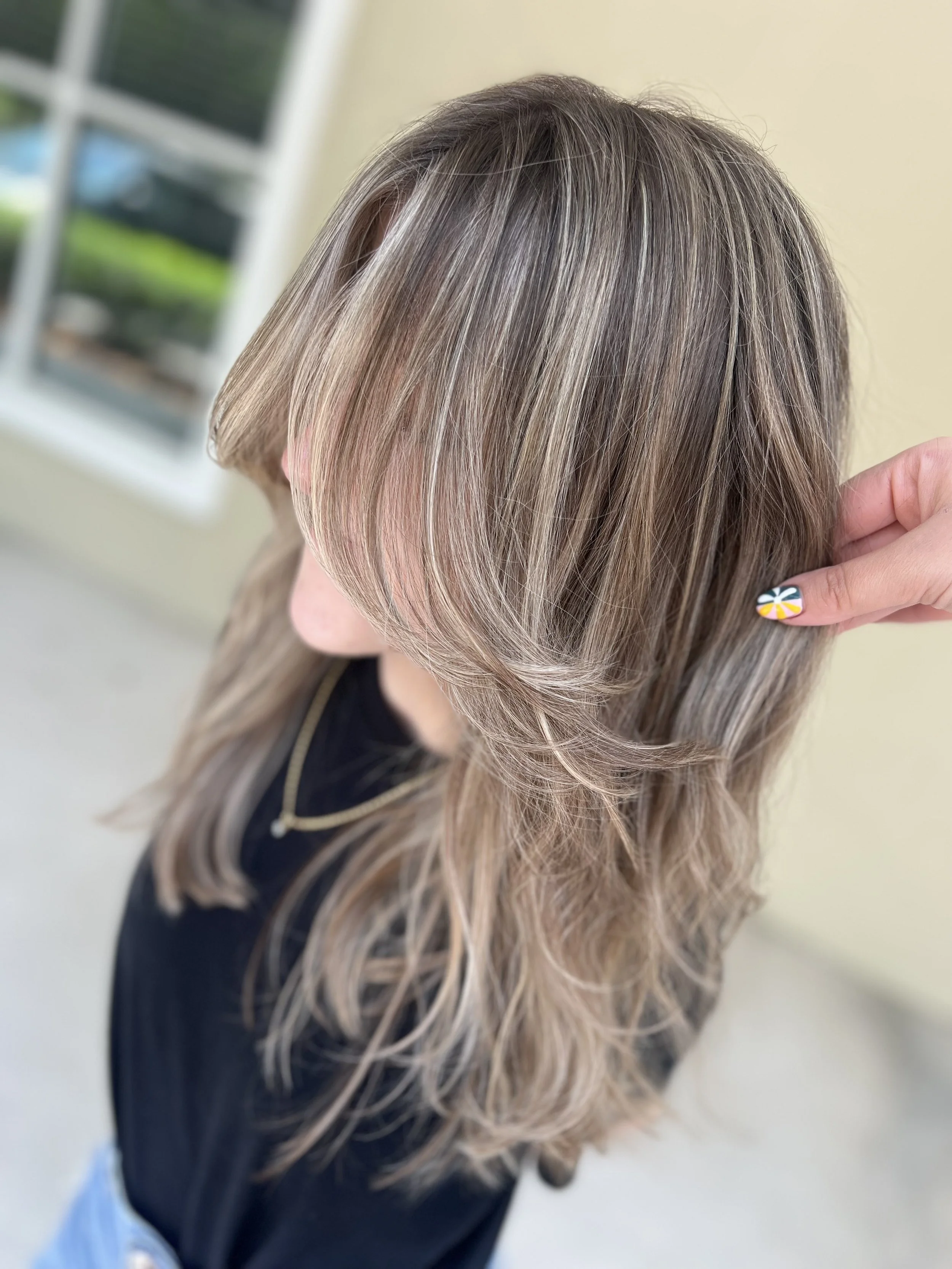 Woman with medium-length, layered blonde hair with subtle highlights, holding a strand with her hand, wearing a black top and a gold necklace, standing near a window.