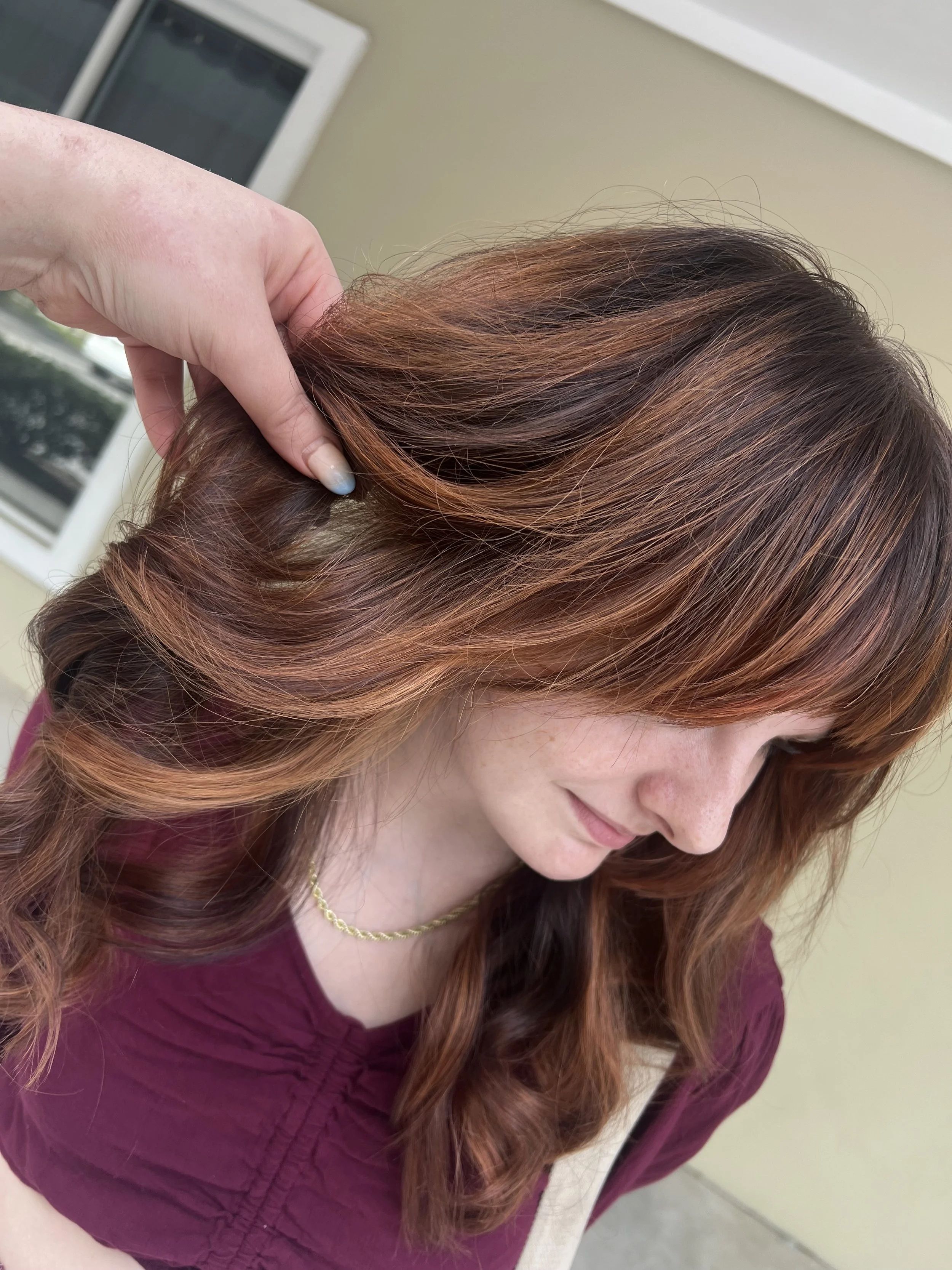 A woman with long, wavy red hair is having her hair styled or examined at a salon. She is smiling and wearing a burgundy top and a gold chain necklace.