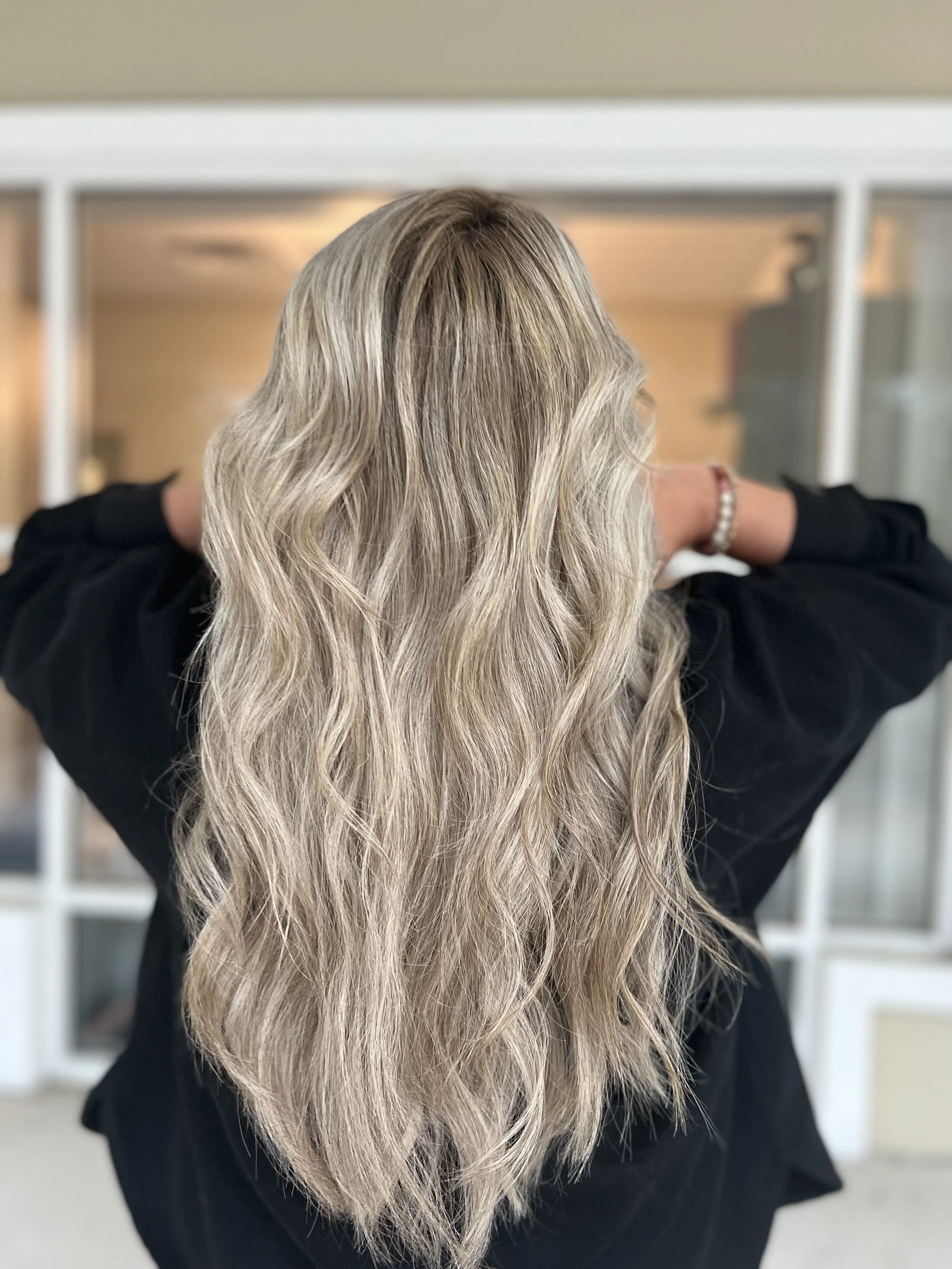 Back view of a woman with long, wavy blonde hair, wearing a black top and a beaded bracelet, standing indoors.