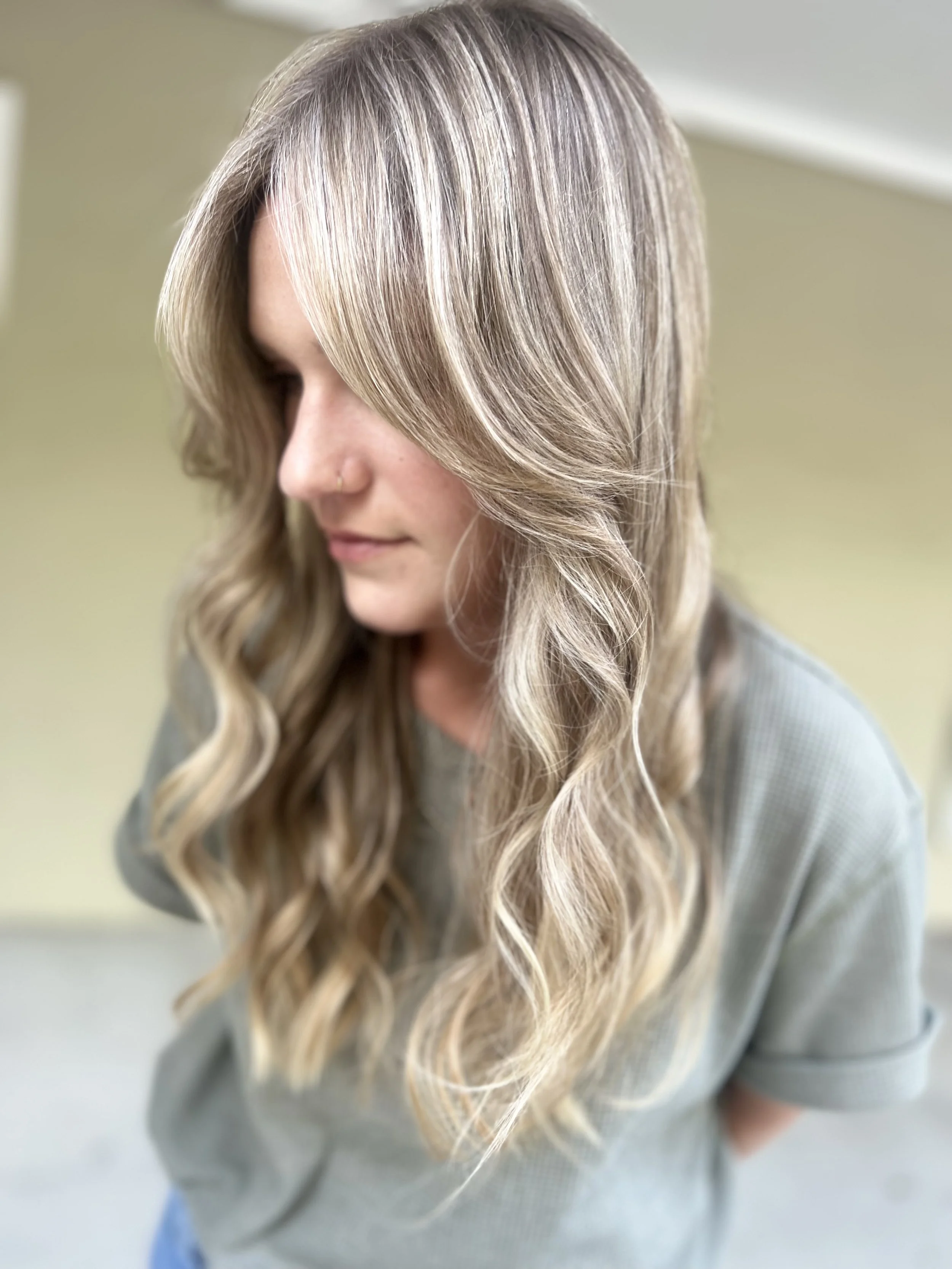 A woman with long, wavy blonde hair and a nose piercing looking down.