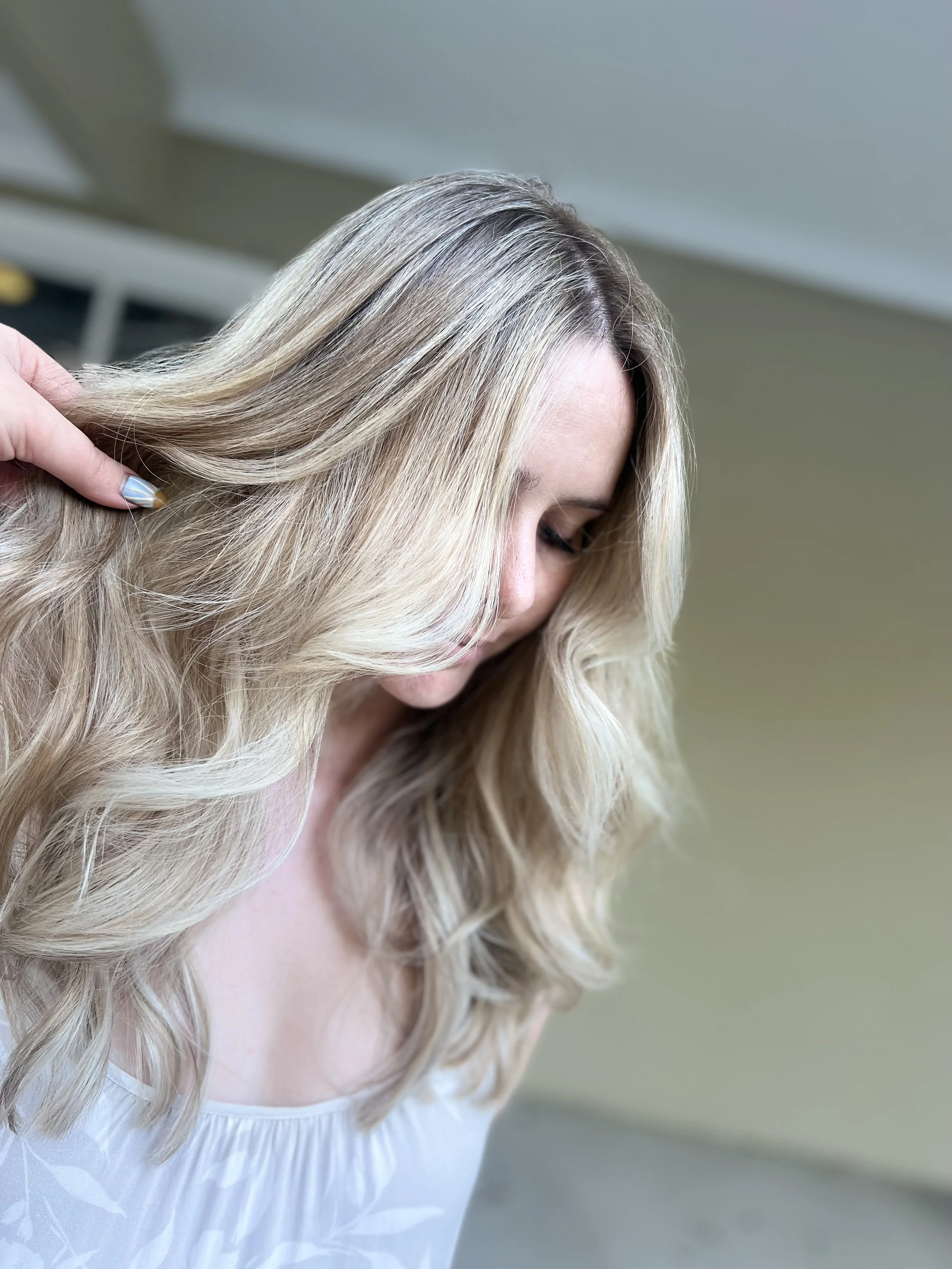 A woman with long, wavy blonde hair looking down, with someone holding a strand of her hair.