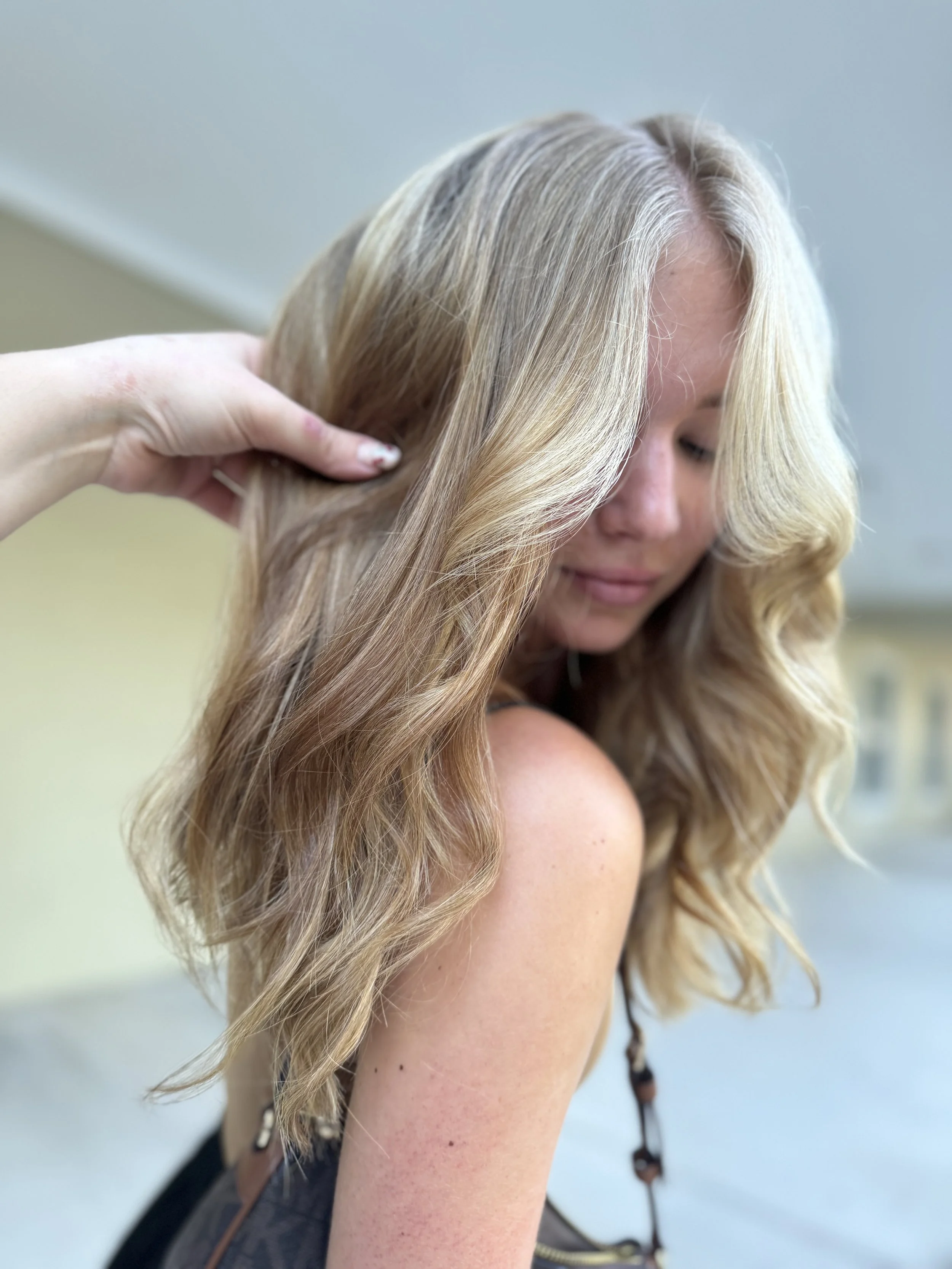 A woman with long, blonde, wavy hair is getting her hair styled by someone whose hand is visible, holding a section of her hair. The woman has her eyes closed and is smiling softly.