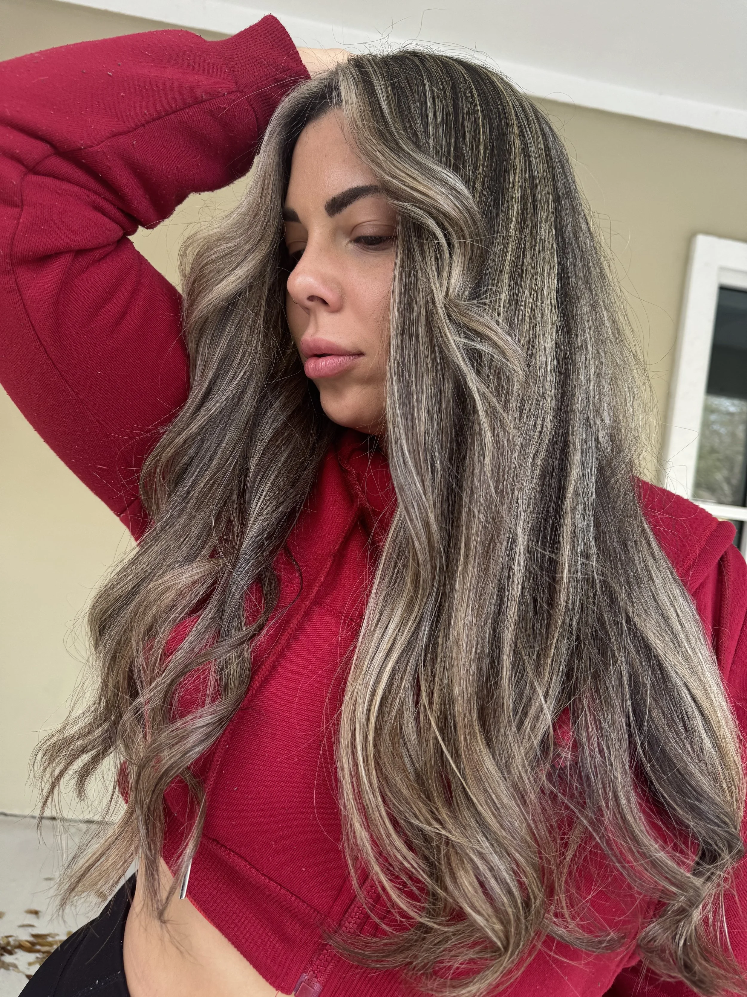 Close-up of a woman with long, wavy, highlighted hair, wearing a red hoodie, standing indoors near a window.