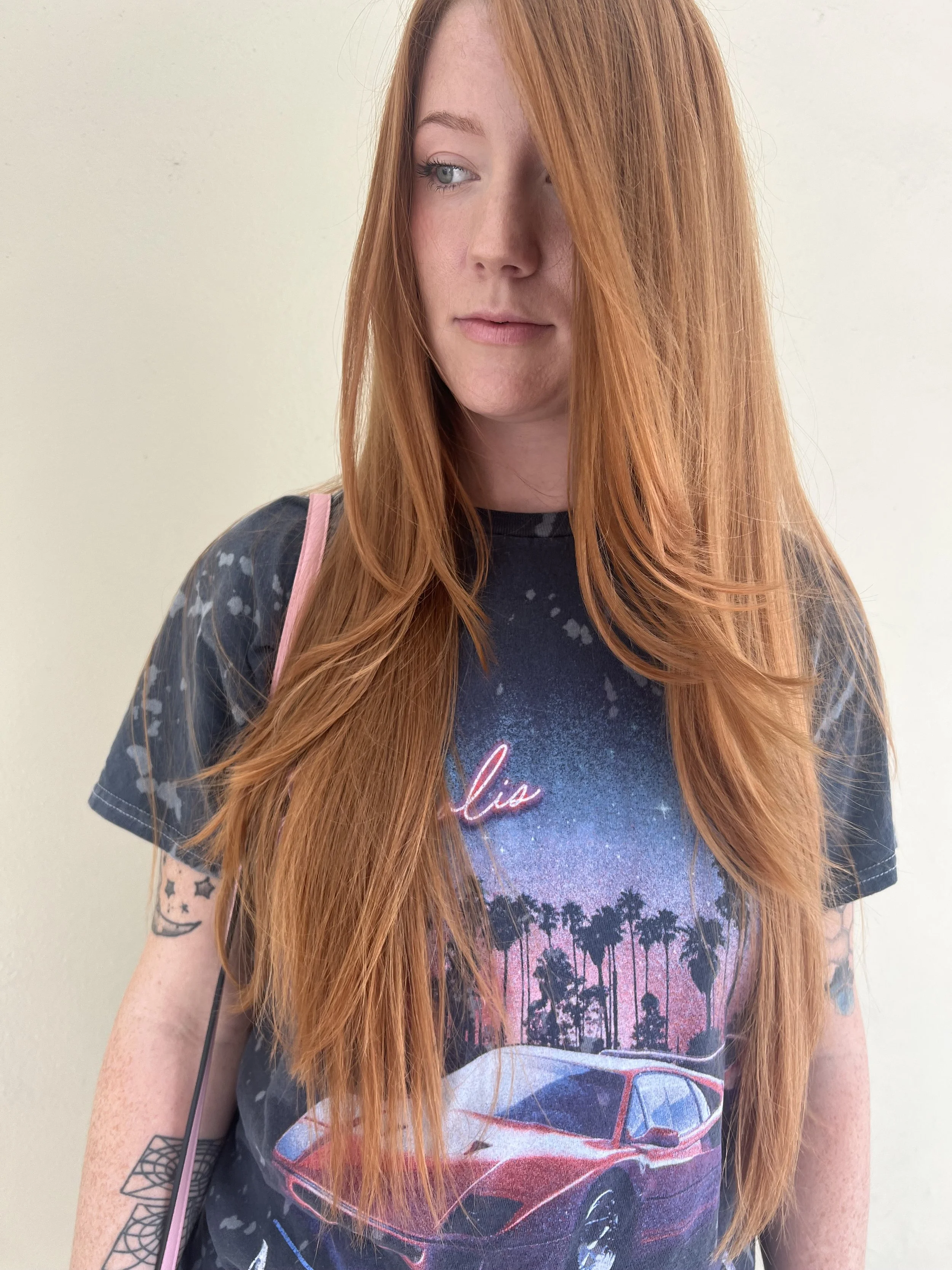 A young woman with long red hair, wearing a black T-shirt with a retro car and palm trees graphic, and small tattoos on her arms, standing in front of a plain light-colored wall.
