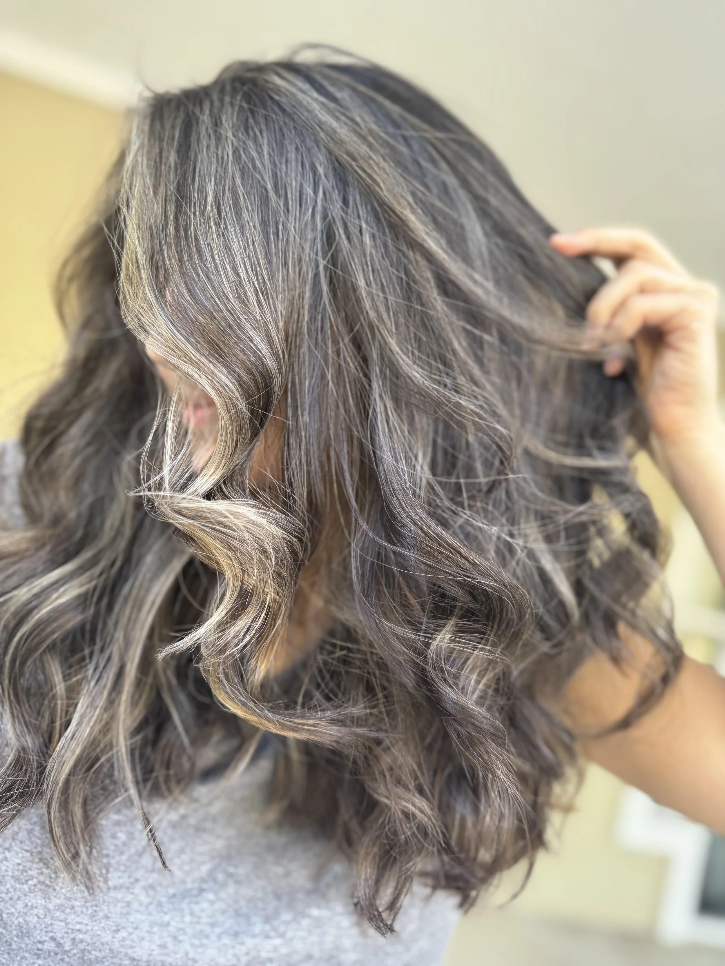 Woman with shoulder-length wavy hair featuring highlights and lowlights, touching her hair with her hand.