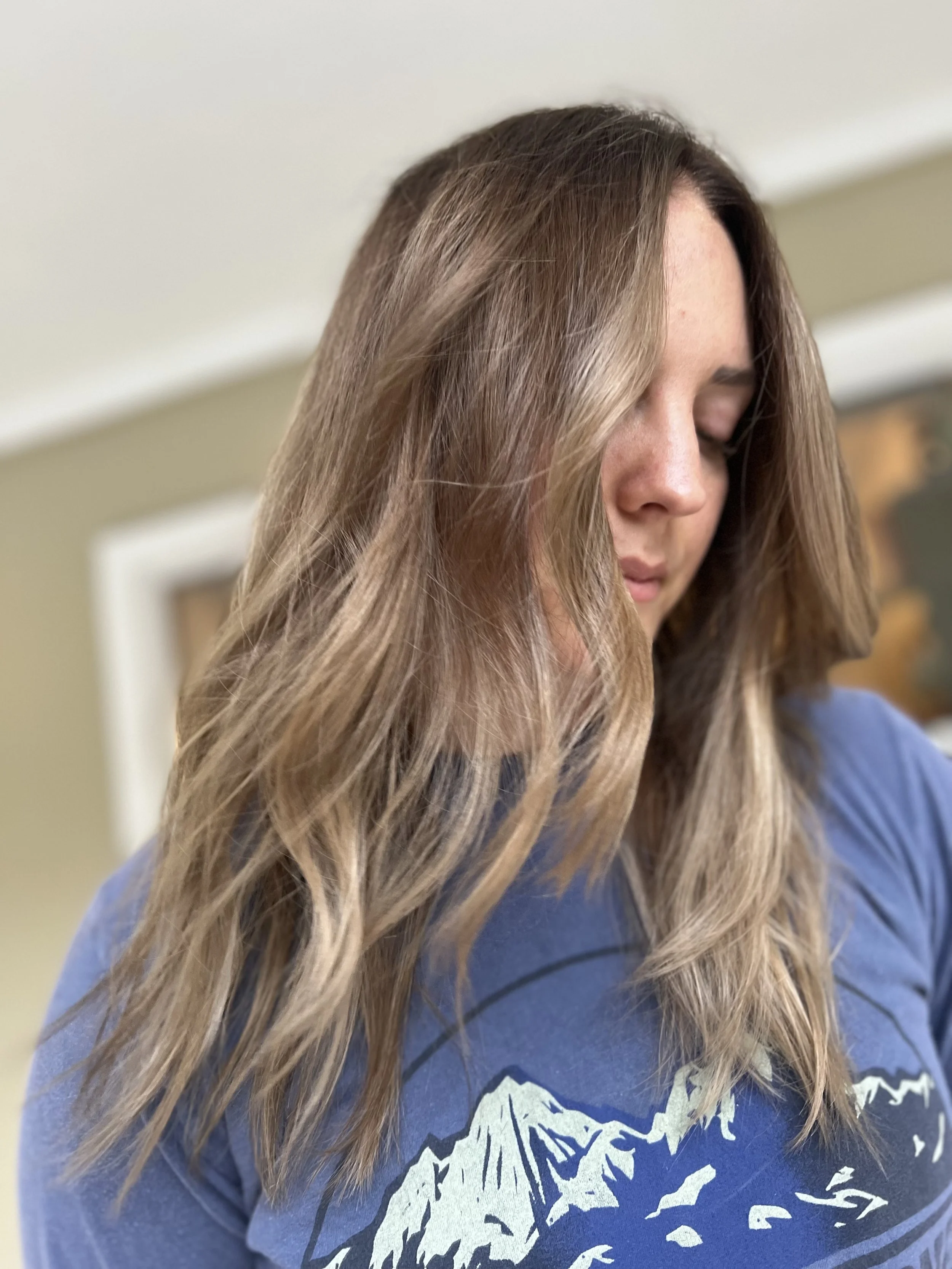 Close-up of a woman with wavy, light brown hair and fair skin, wearing a blue T-shirt with a mountain graphic, looking downward with eyes closed, in an indoor setting.