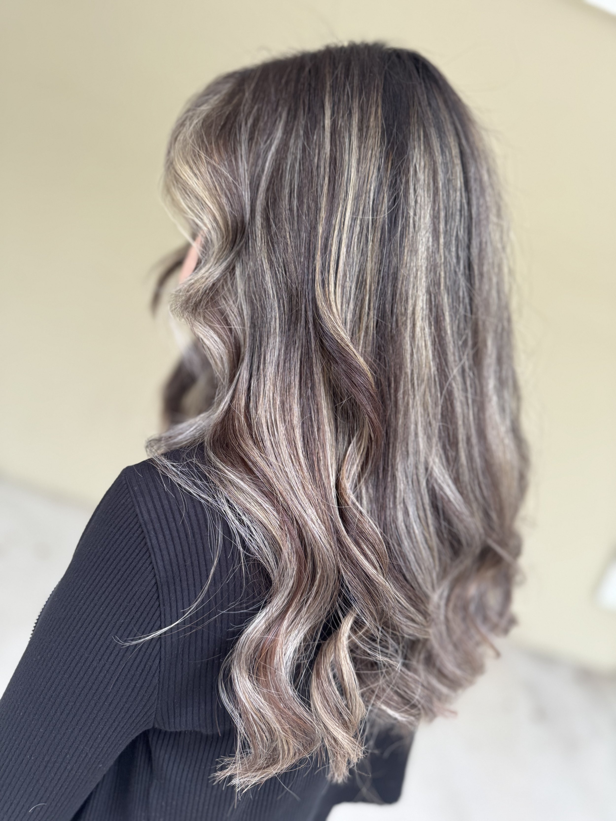 Woman with shoulder-length, wavy hair with blonde highlights and darker roots, wearing a black long-sleeve top.