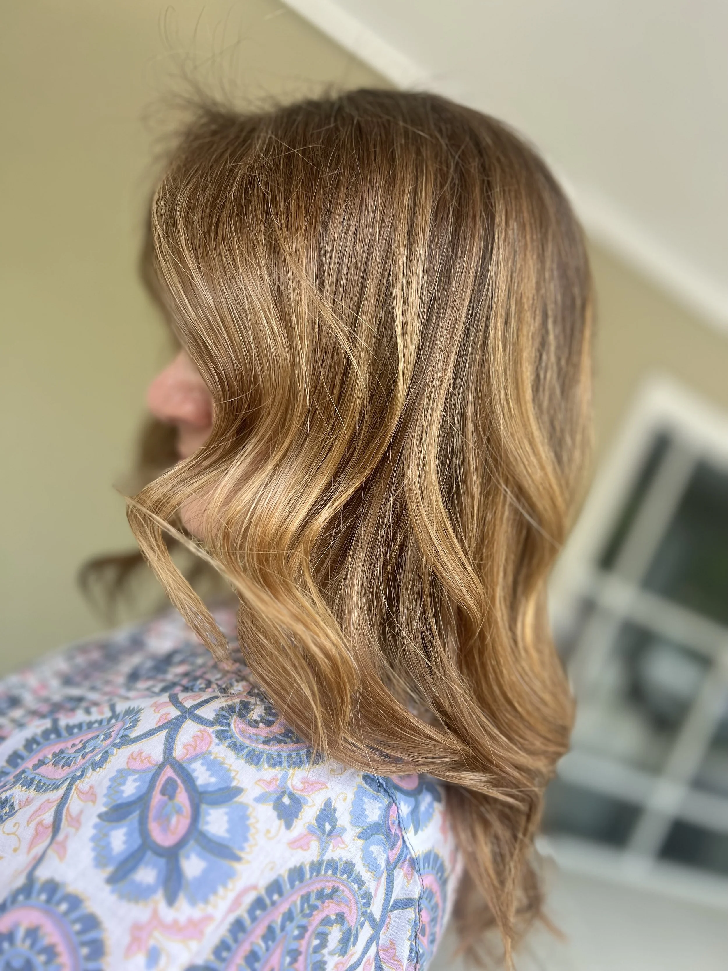 Close-up side view of a woman with wavy, shoulder-length, reddish-brown hair. She is wearing a patterned top with blue and pink designs, and there is a blurred background with a window.