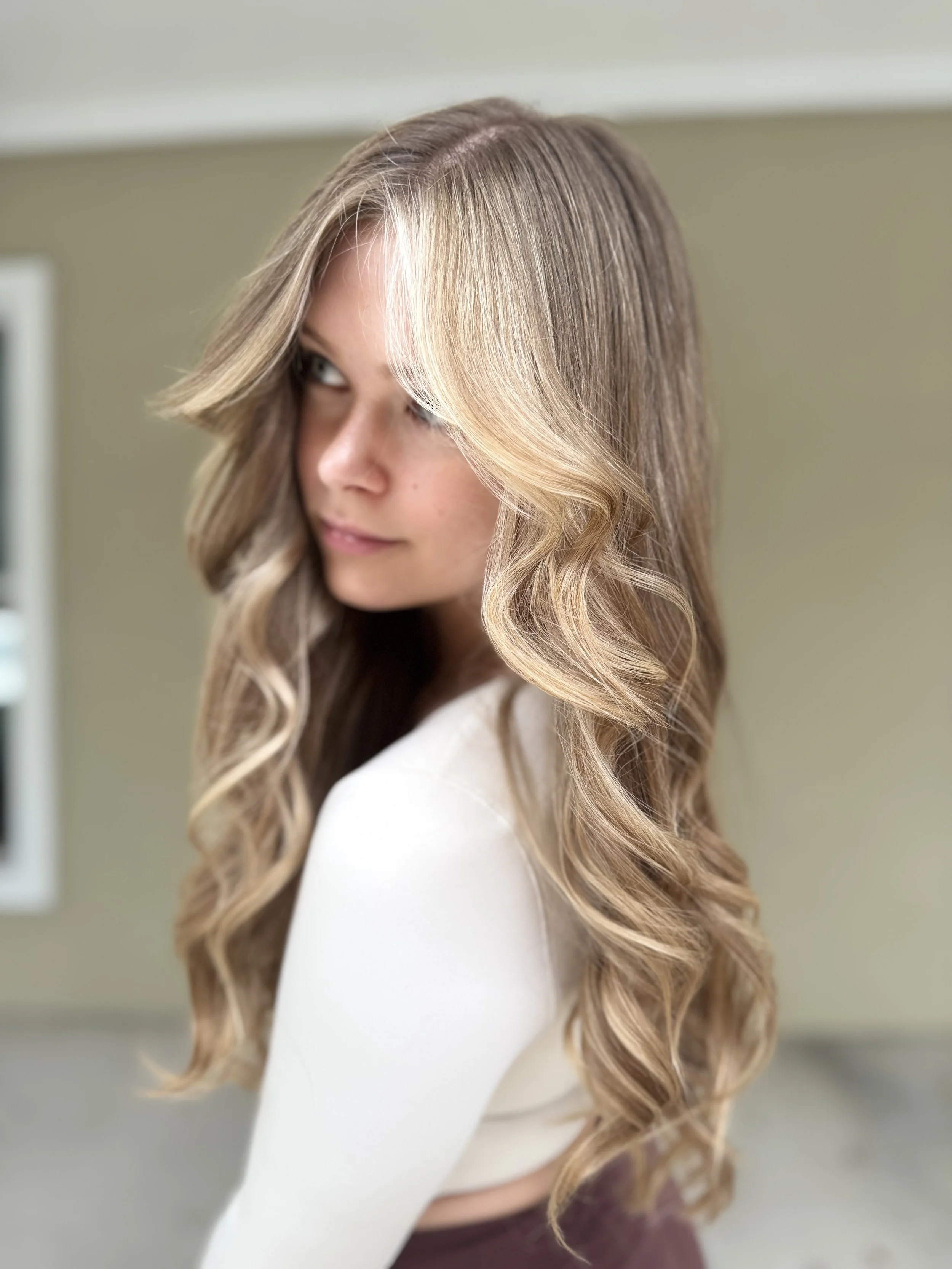 A young woman with long, wavy blonde hair and fair skin, looking sideways with a slight smile, indoors with soft lighting.
