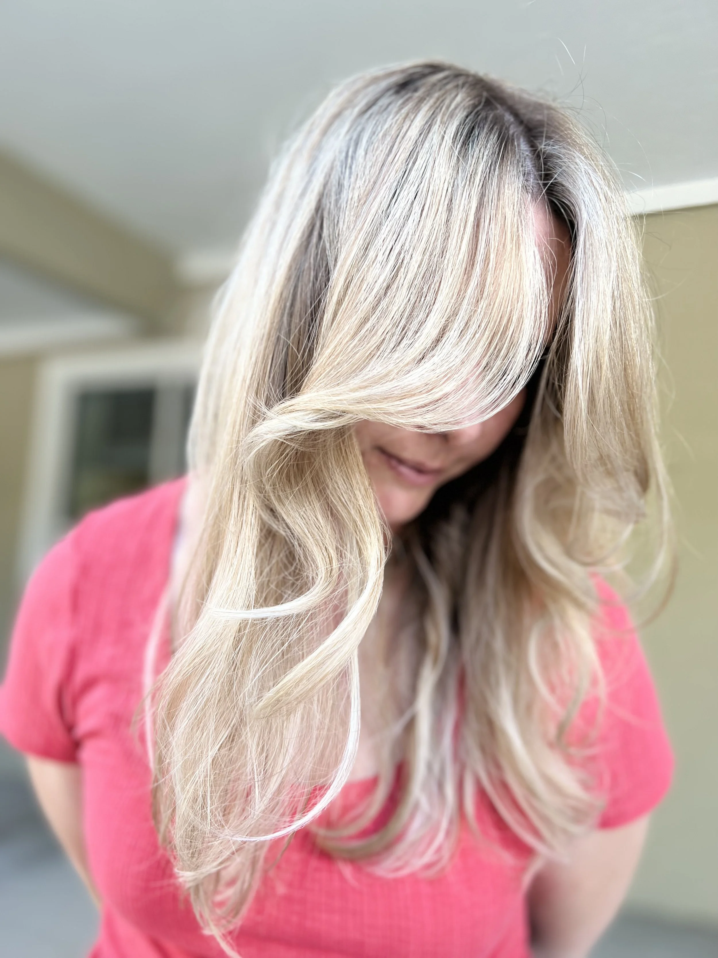 A woman with long, wavy blonde hair wearing a pink top, looking down with her hair covering part of her face.