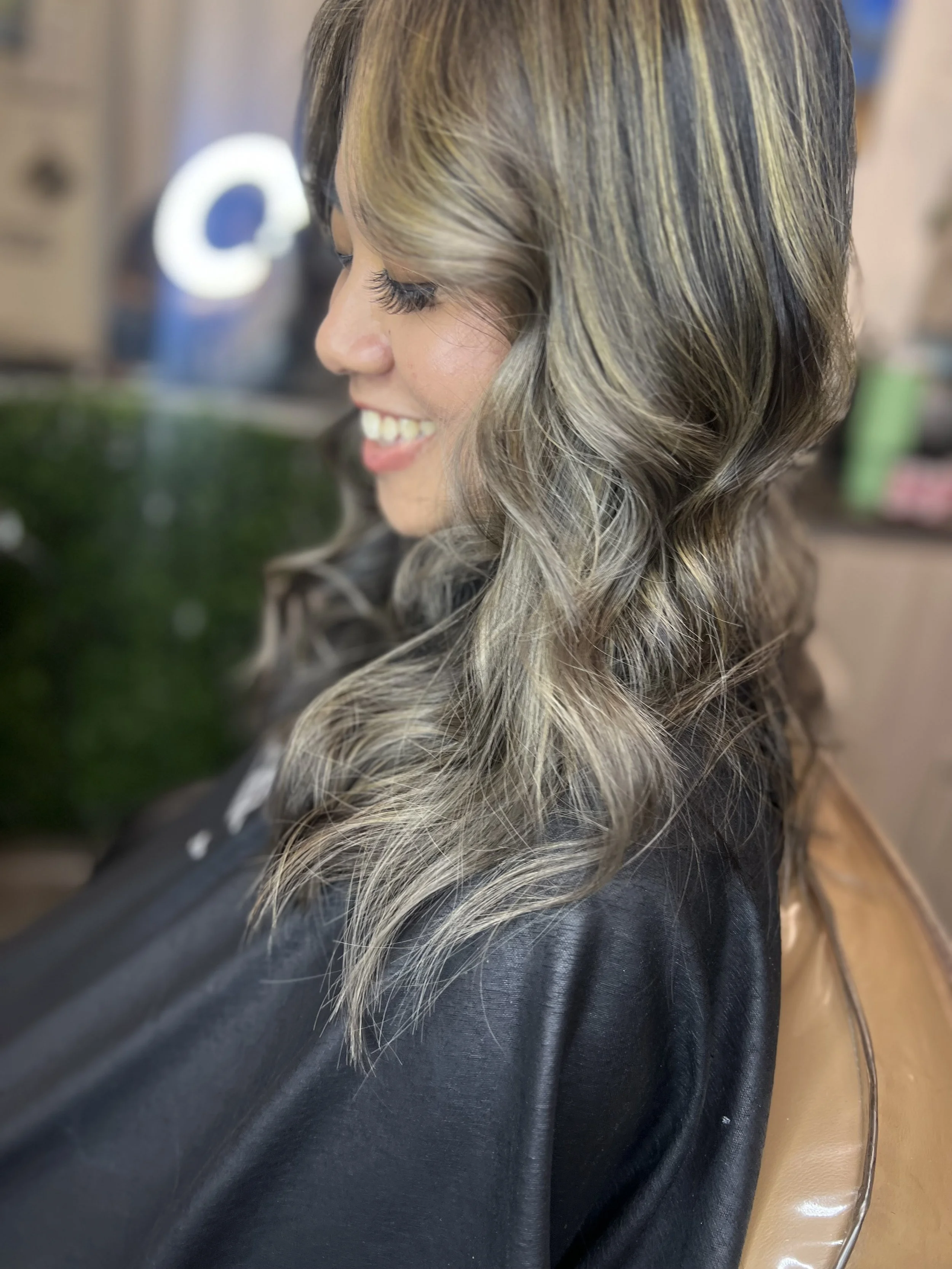 Side view of a smiling woman with styled wavy blonde and brown hair, wearing a black cape, sitting in a salon chair.