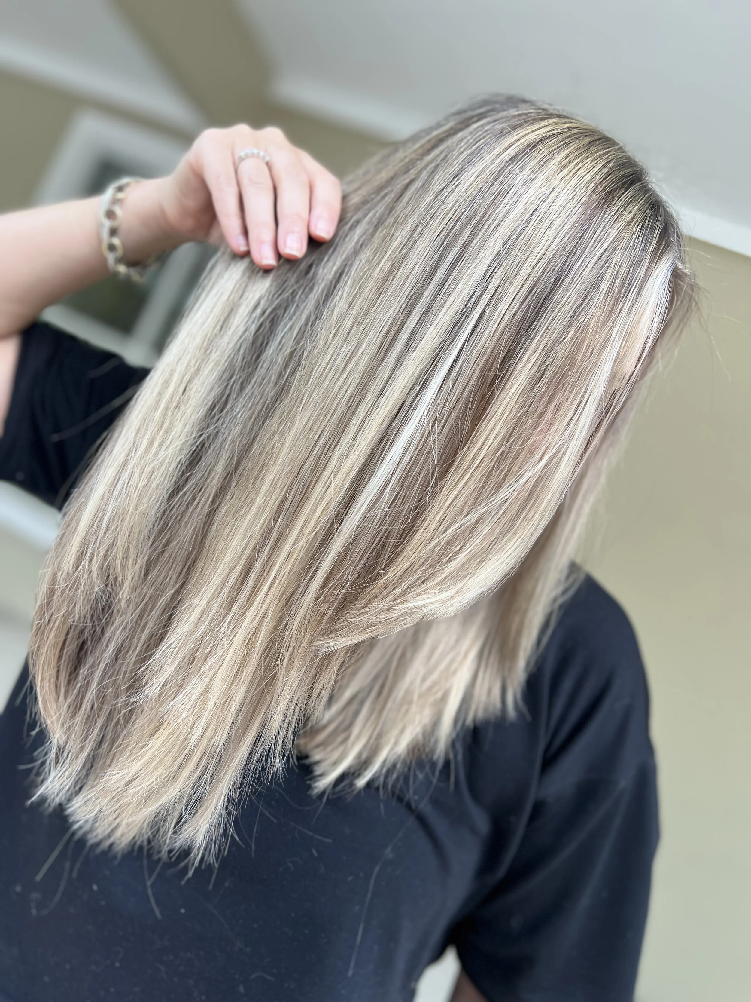 Close-up of a woman with straight, shoulder-length blonde hair, wearing a black shirt and a silver bracelet, touching her hair with her hand.