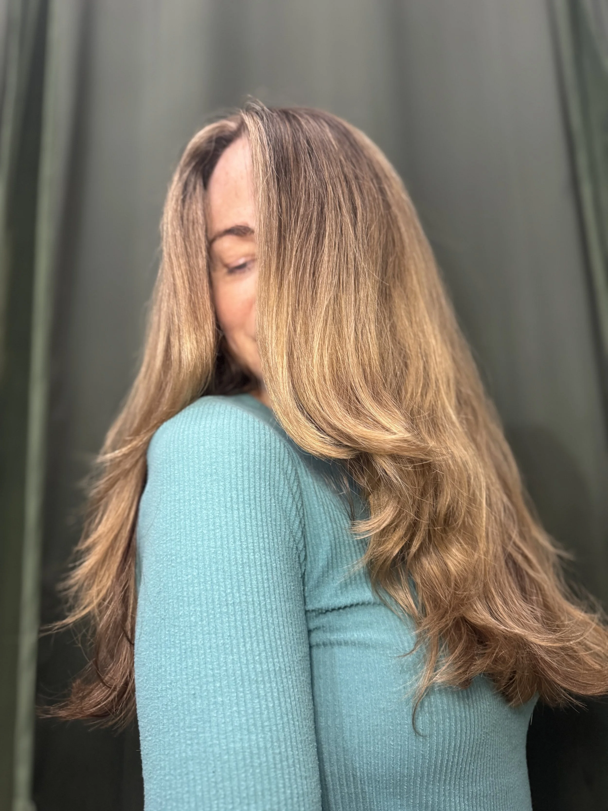 Woman with long, wavy, brown hair wearing a teal long-sleeve top, standing in front of a dark background.