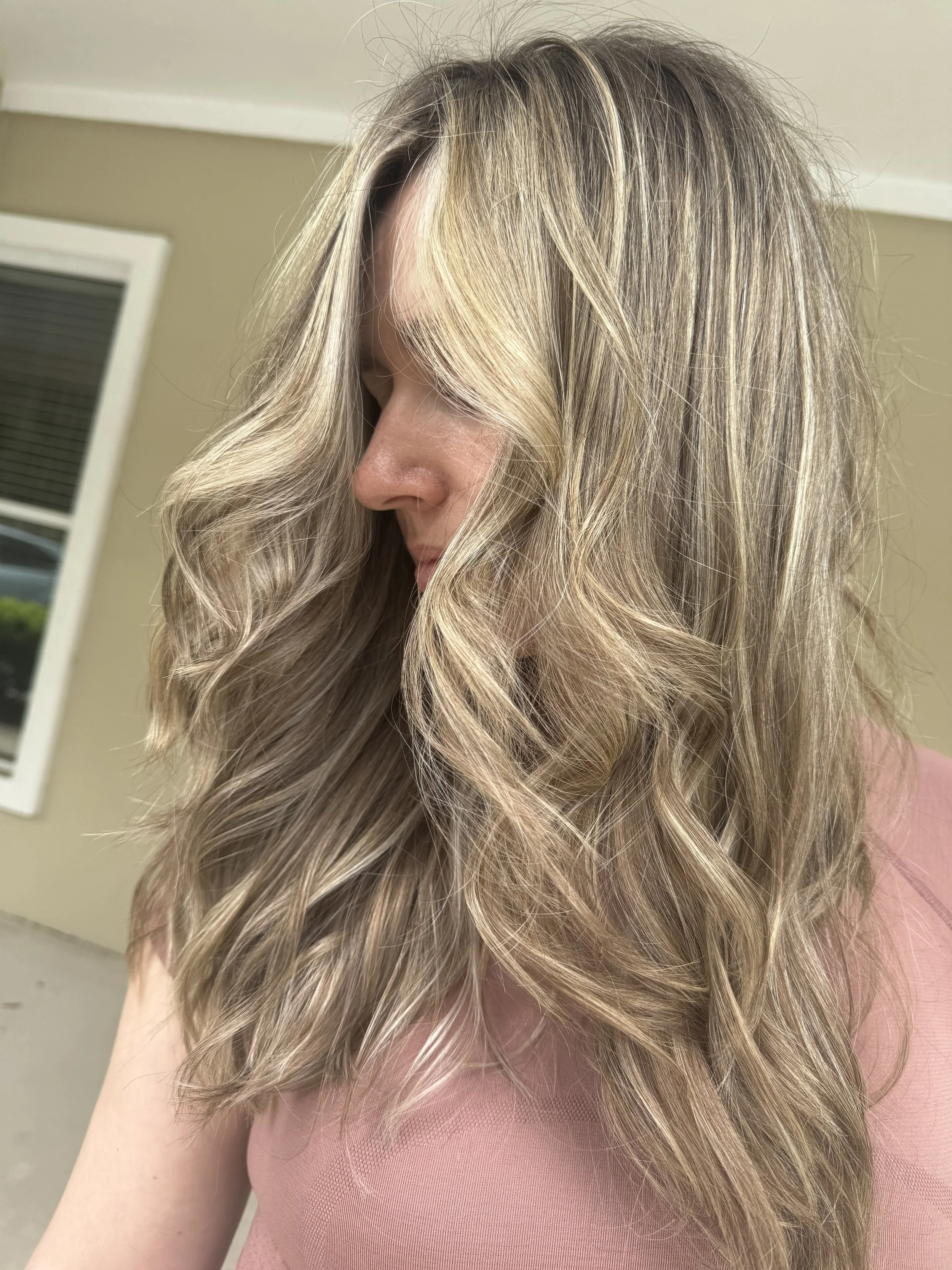 Woman with long, wavy blonde hair, wearing a pink top, standing indoors near a window.