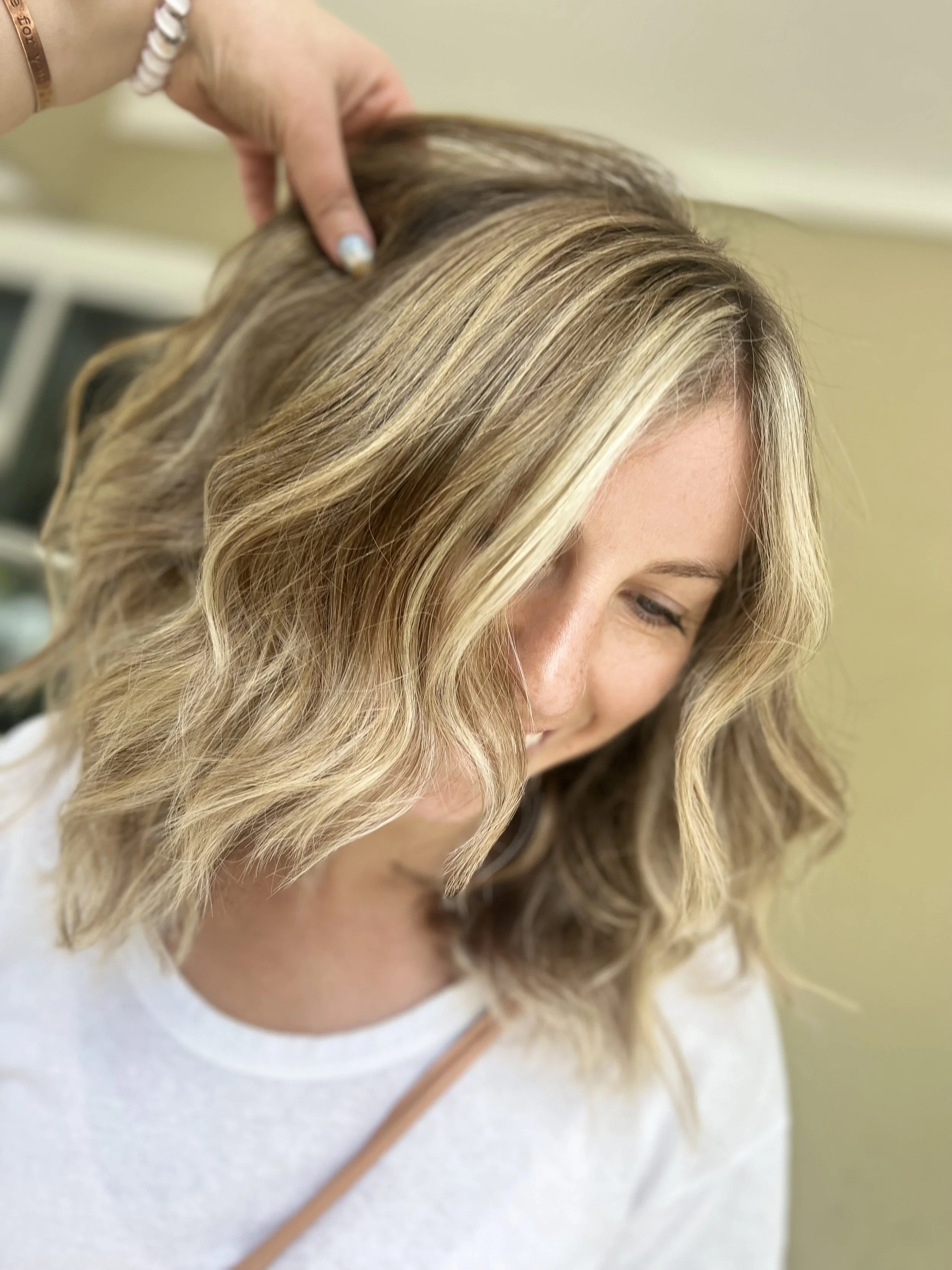 A woman smiling with shoulder-length blonde hair with soft waves, having her hair styled by a person with a hand on her head, in a salon or similar setting.