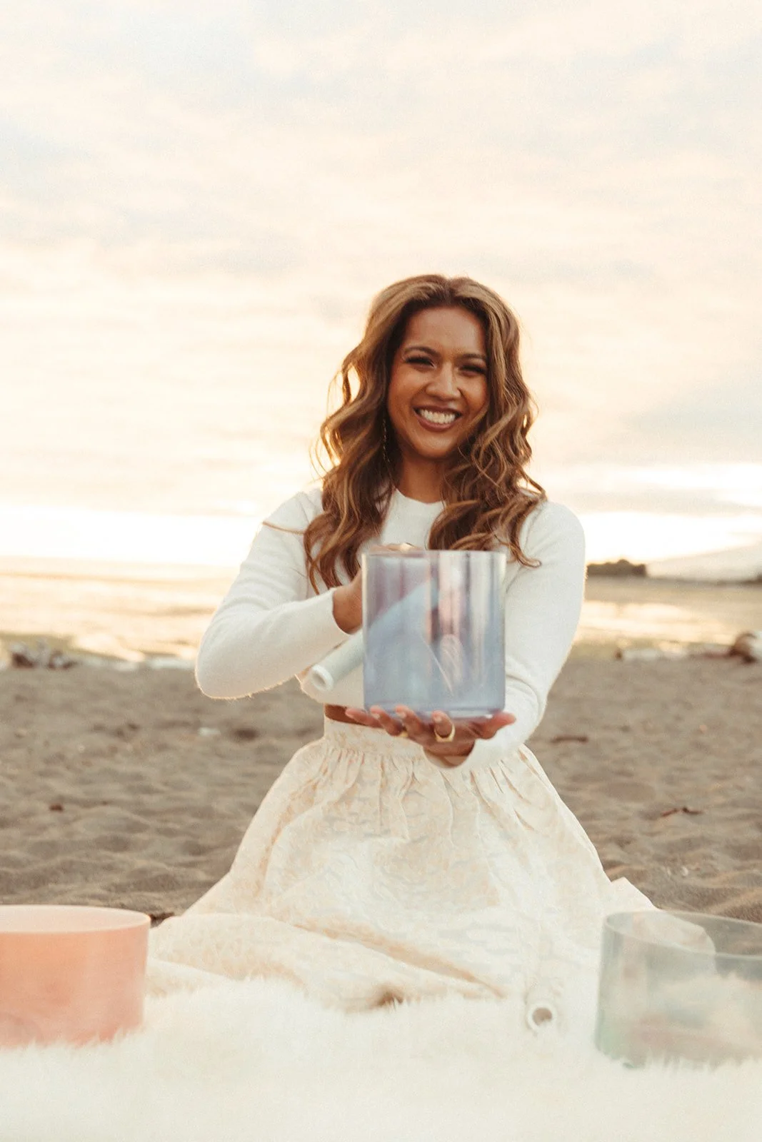 A woman sitting on the beach at sunset, smiling and holding a crystal singing bowl.