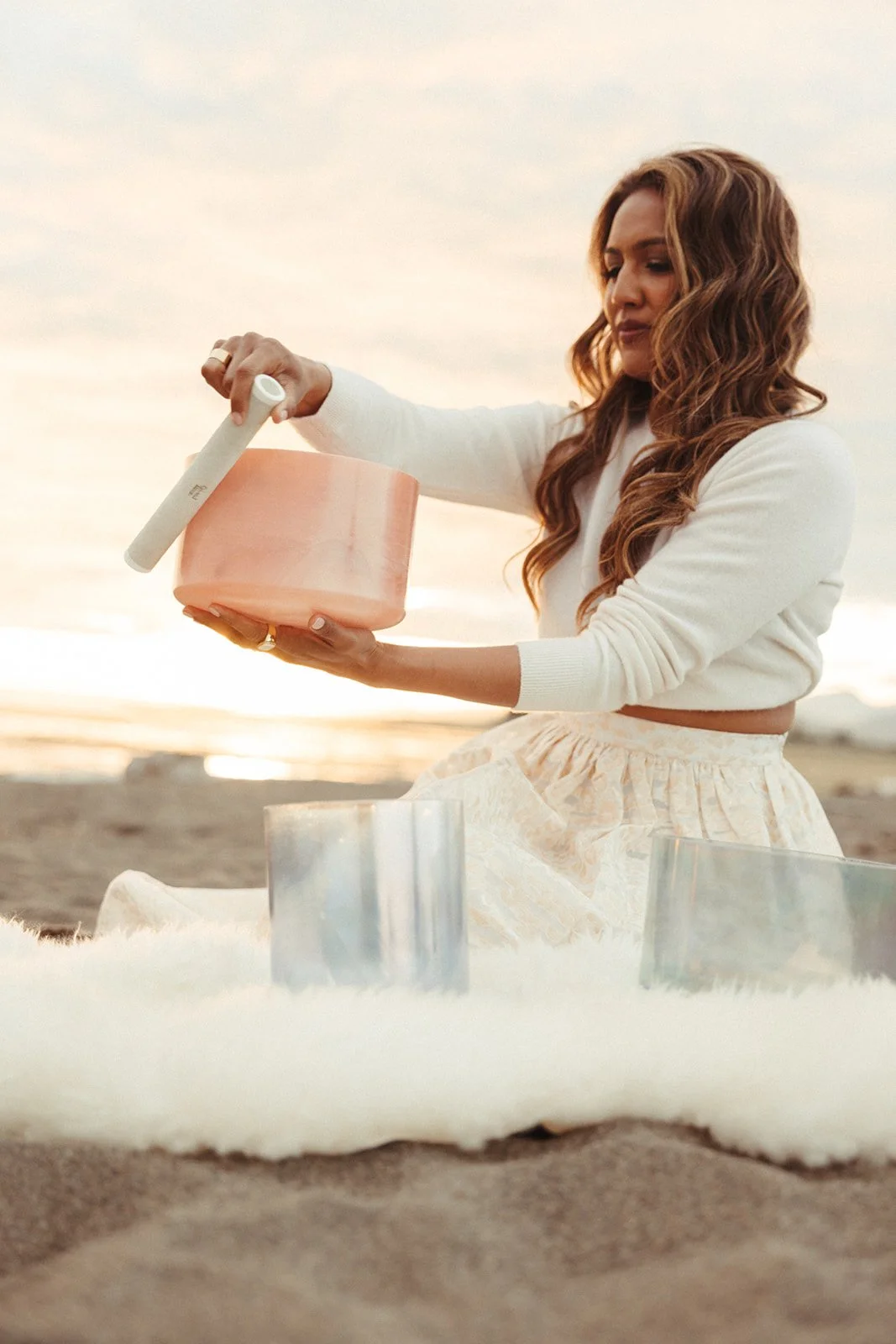 Henrietta Devine of Sound Space Portal A woman participates in a spiritual or meditative ceremony on the beach at sunset, holding a pink alchemy crystal tone singing bowl with ritual objects inside.