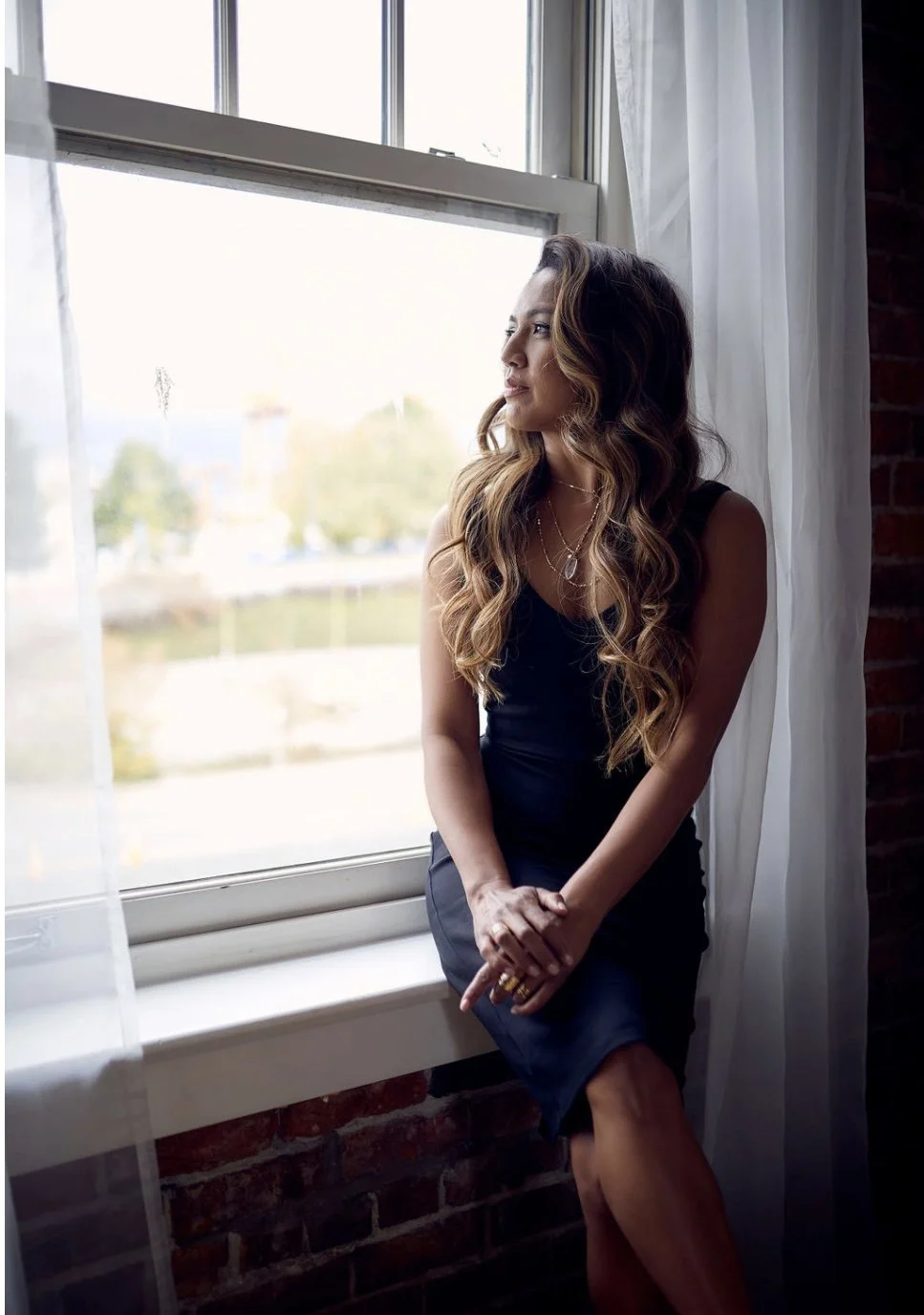 A woman with wavy hair in a black dress and layered jewelry looking out a window with white curtains, interior studio setting with brick wall.