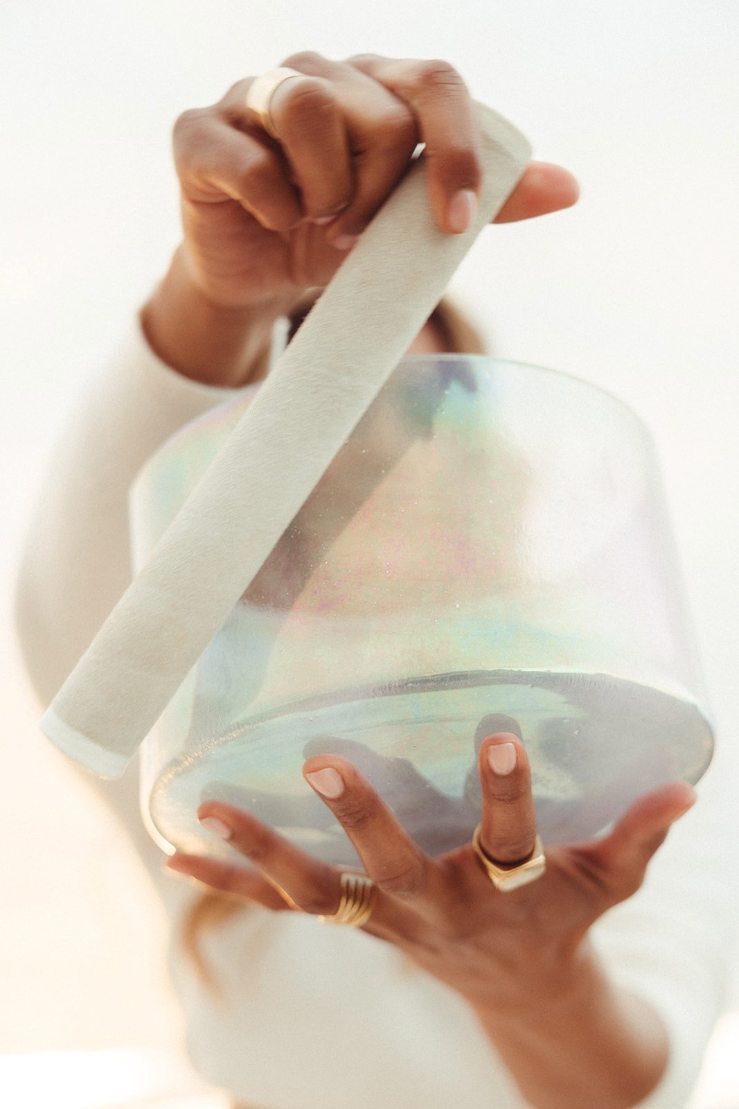 A person holding a clear, iridescent alchemy crystal singing bowl with one hand and a white baton with the other hand, against a plain light background.