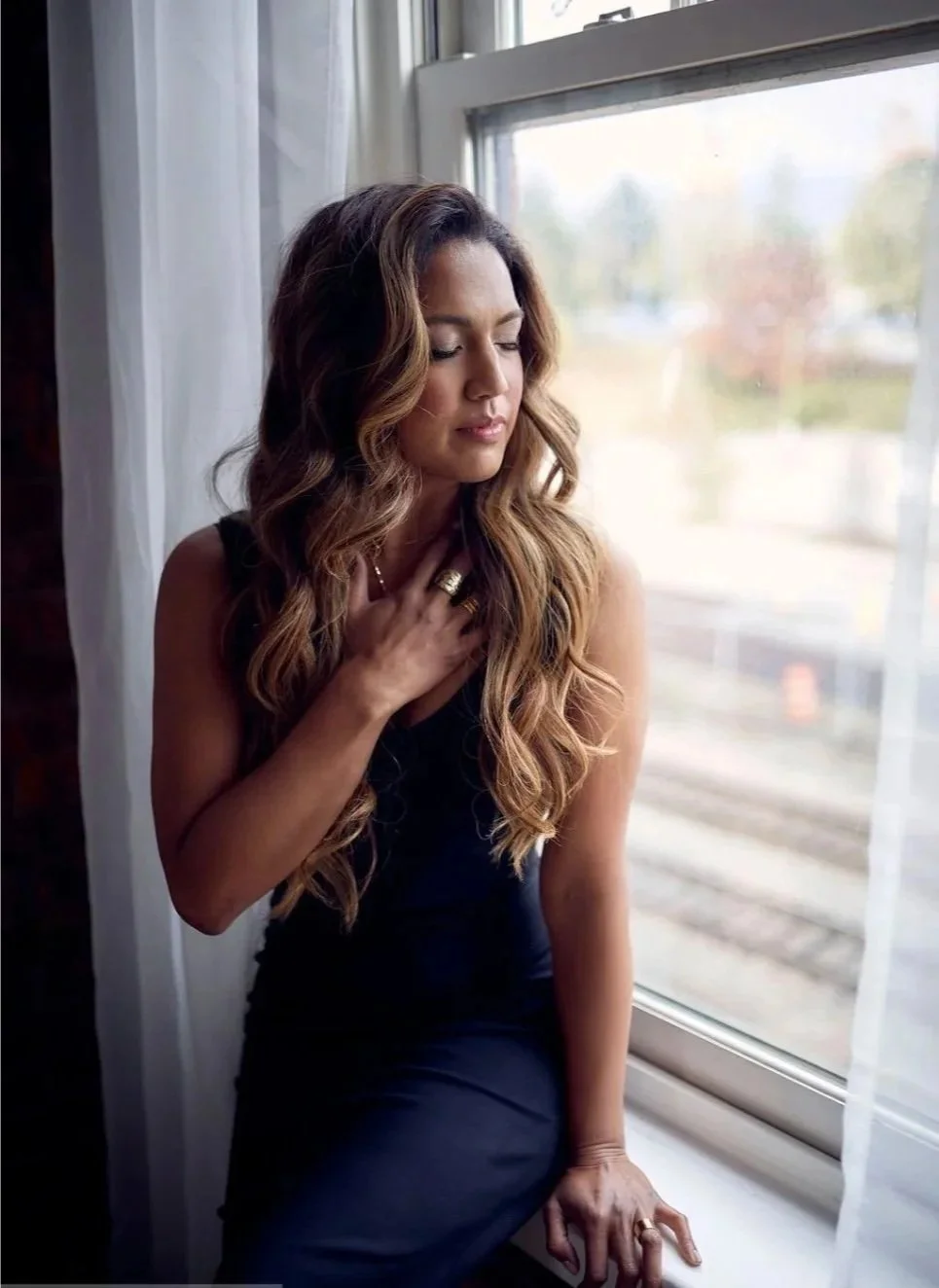 Woman with long wavy hair wearing a black dress, standing by a window with sheer curtains, looking contemplative with eyes closed and hand on chest. Maestra Henrietta Devine, Vancouver frequency architect and sound journey practitioner