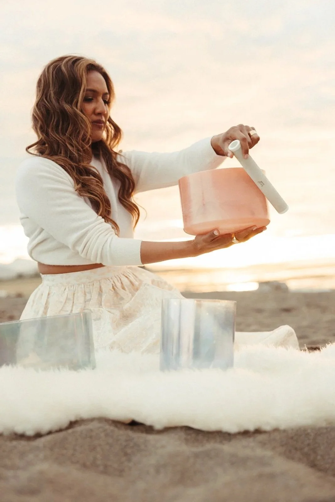 A woman with long, wavy hair stands on a beach during sunset, holding a pink bowl and a white mallet, surrounded by clear crystal bowls and a white furry blanket.