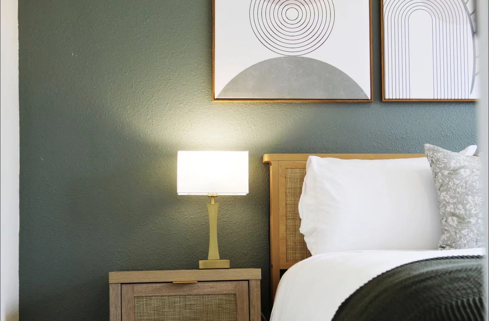 A bedroom with a green wall, a wooden bed with a white pillow and patterned pillow, a wooden nightstand, a yellow lamp, and two abstract framed artworks on the wall.