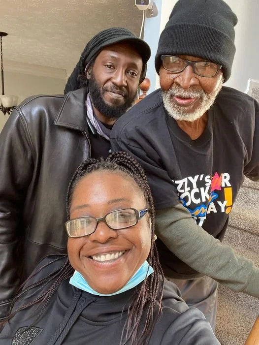 Three people smiling for a selfie indoors. The woman in the foreground is wearing glasses, a black jacket, and a face mask under her chin. The man on the right is wearing glasses, a black beanie, and a T-shirt with colorful text. The man on the left is wearing a black jacket and has a short beard.