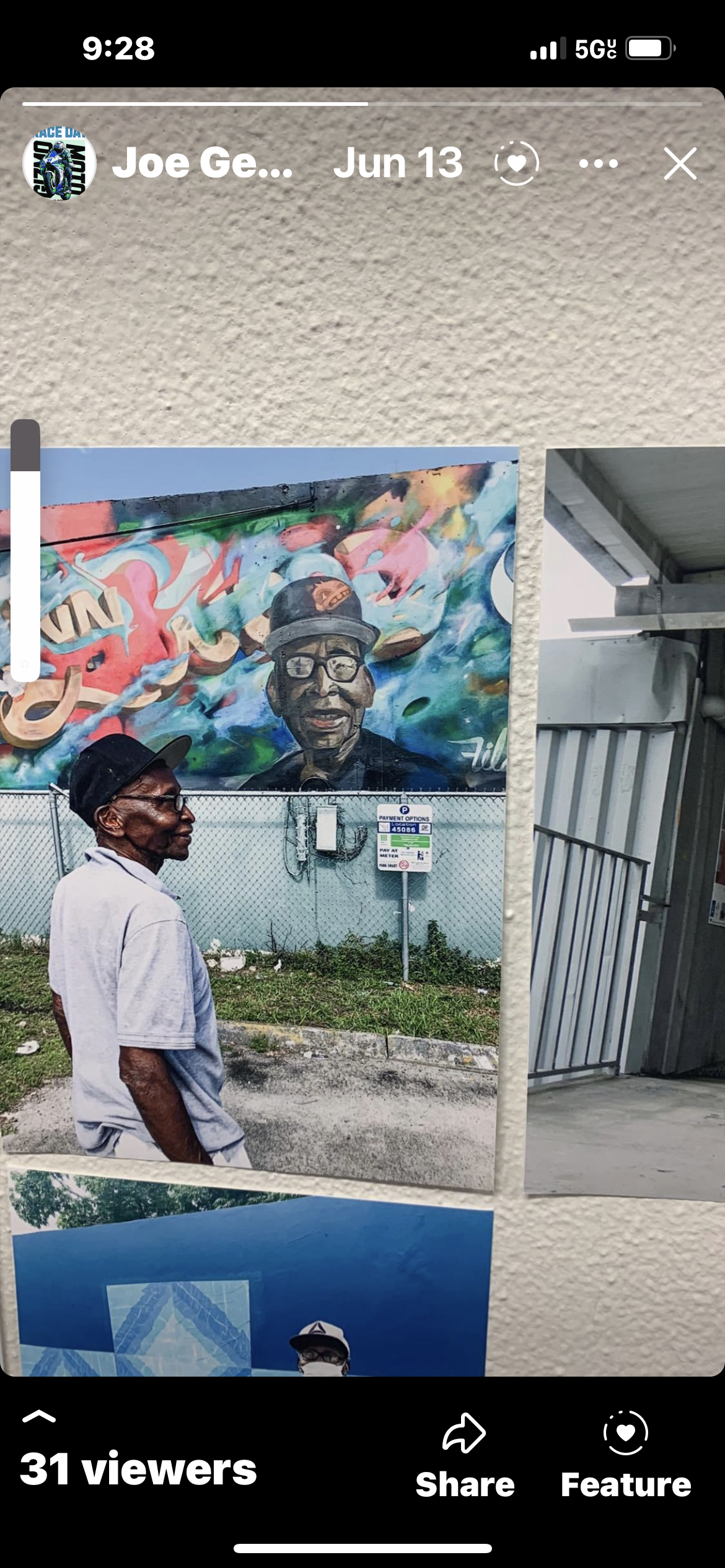 A man with glasses and a black hat standing in front of a colorful mural of a smiling person wearing glasses and a cap, painted on a wall. Another person is partially visible in the lower part of the image.