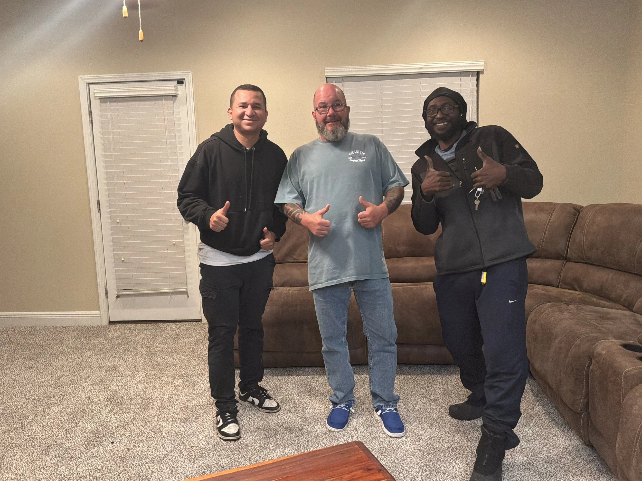 Three men smiling and giving thumbs up in a living room with a beige couch and brown carpet.