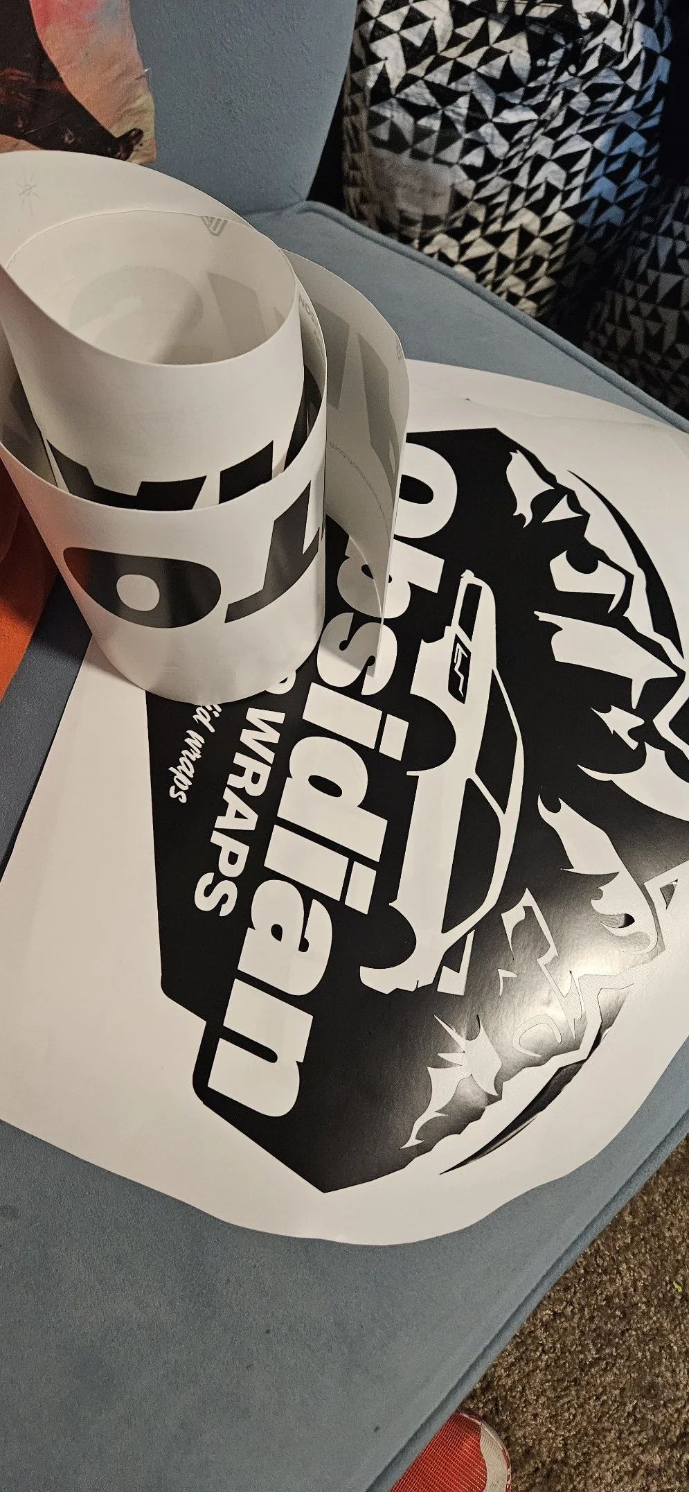 Black and white printed car graphic sticker sheet with the text 'The Best Rides' and a stylized car illustration, partially peeled off, resting on a blue surface with a rolled paper strip and patterned pillows in the background.