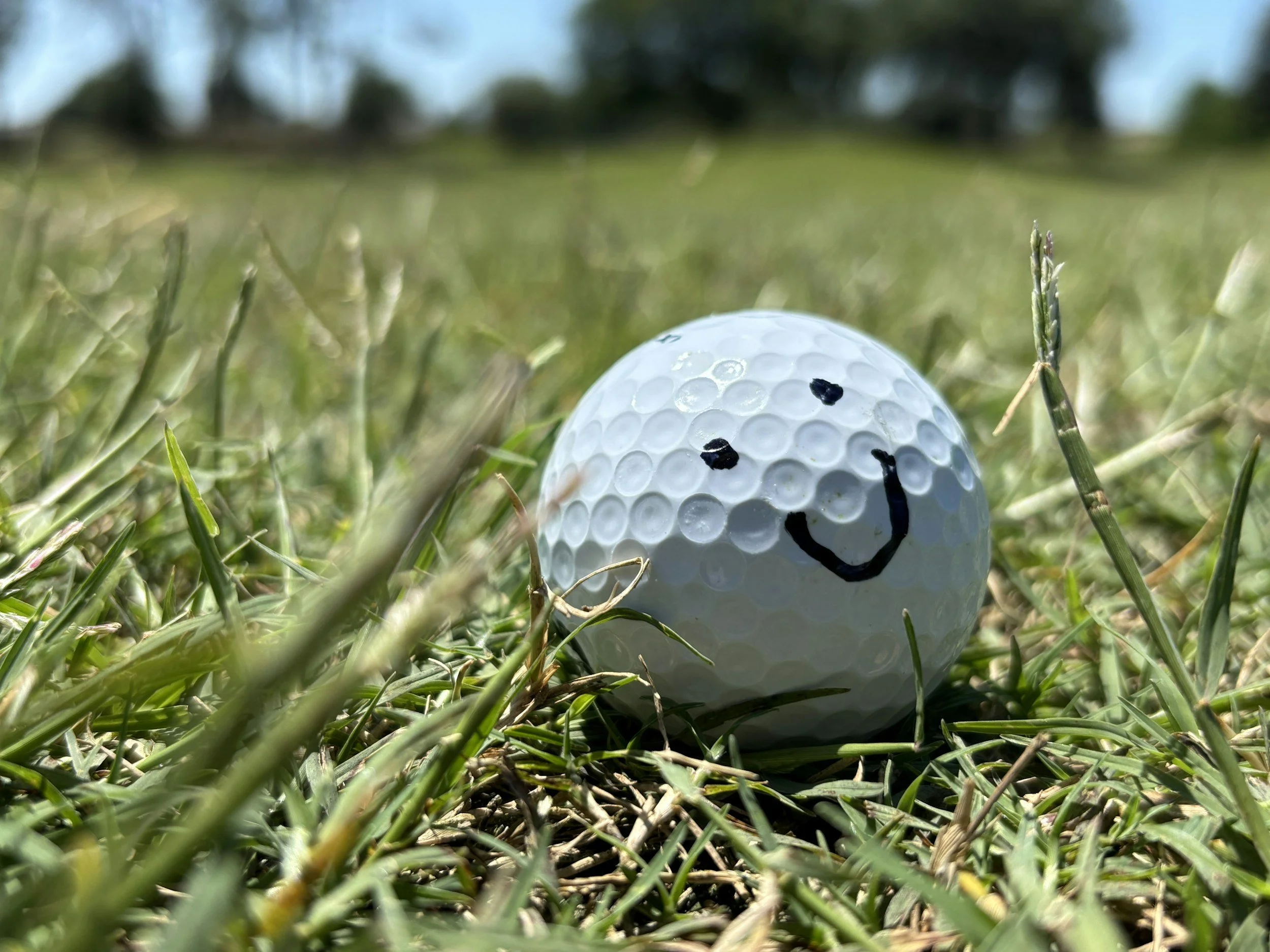 Close-up of a golf ball with a smiley face drawn on it, lying in the grass on a golf course.