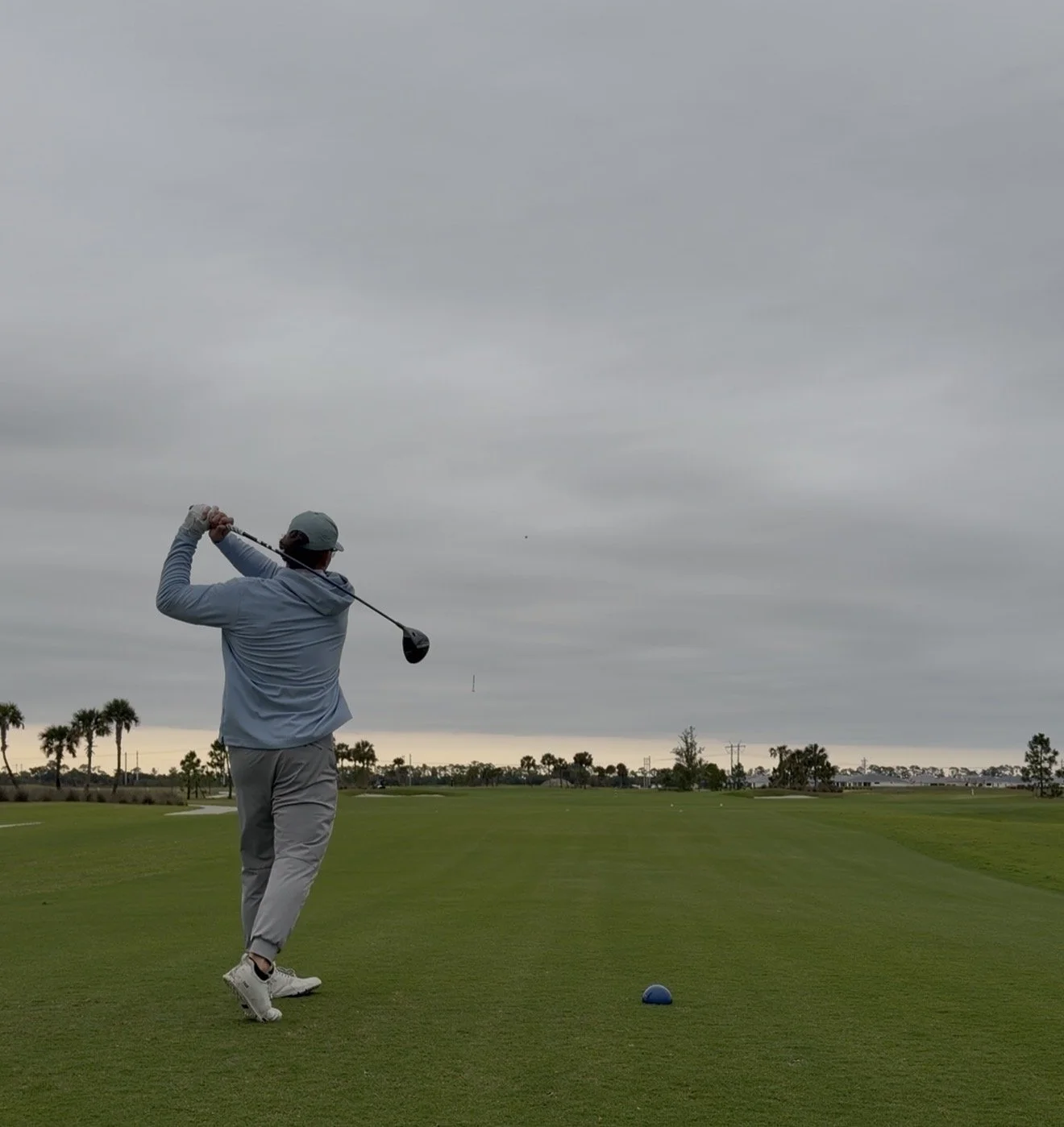 A golfer swings a club on a cloudy day at a golf course with palm trees in the background.