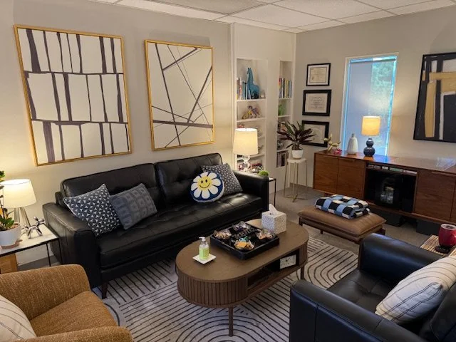 Cozy living room with black sofas, patterned pillows, a wooden coffee table, and wall art. There are lamps, a bookshelf with decorative items and framed certificates, and a sideboard with a table lamp and decor. Natural light comes through a window.