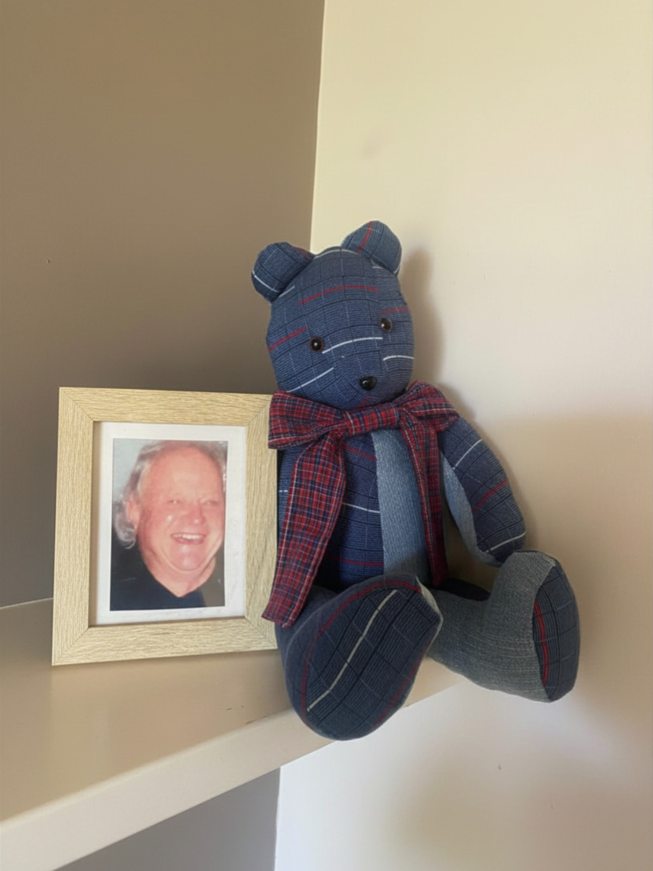 A plaid teddy bear with a red and black bowtie sitting next to a framed photo of an elderly man on a white shelf.