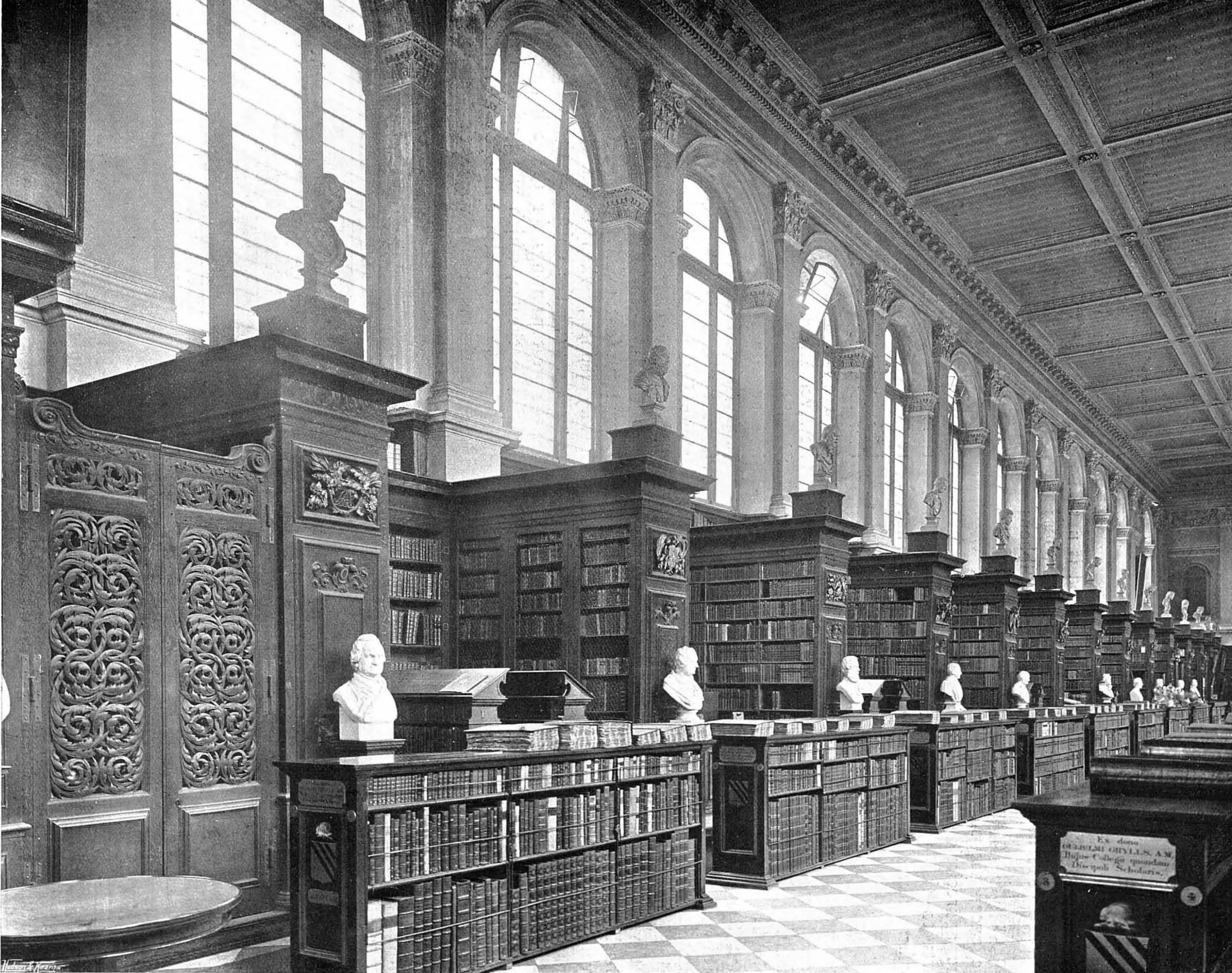 Black and white photograph of a grand library interior with tall arched windows, ornate wooden bookshelves filled with books, marble busts on pedestals, and classical statues. The high ceiling has decorative wooden panels, and the floor is patterned with tiles.