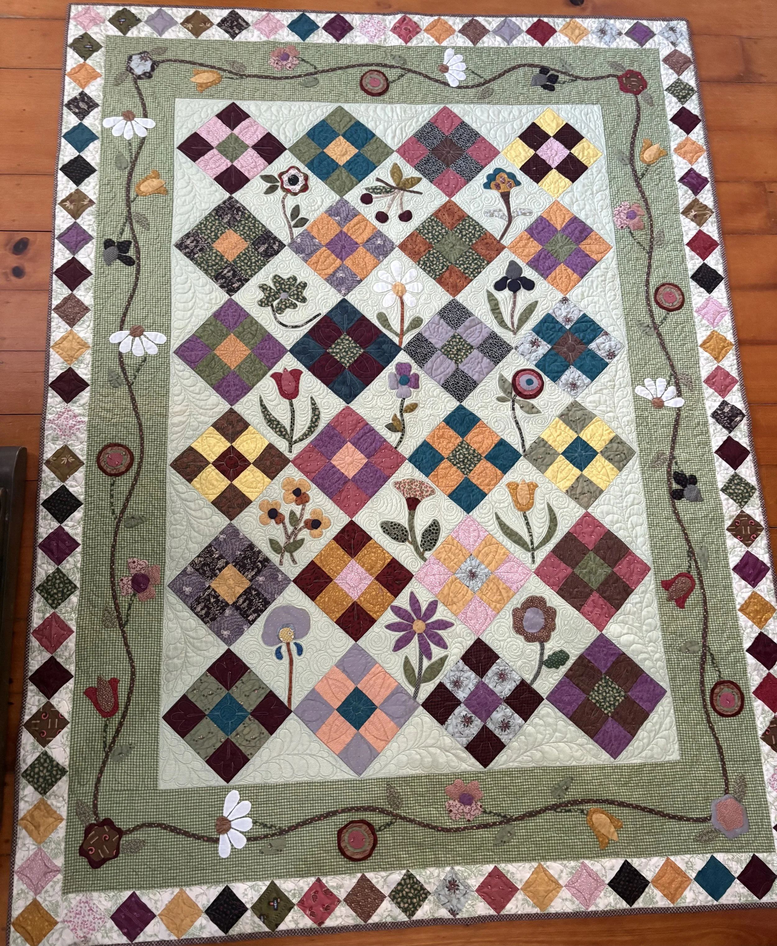 Colorful quilt with floral embroidery and checkerboard pattern, featuring flowers, cherries, and decorative border on a wooden floor.