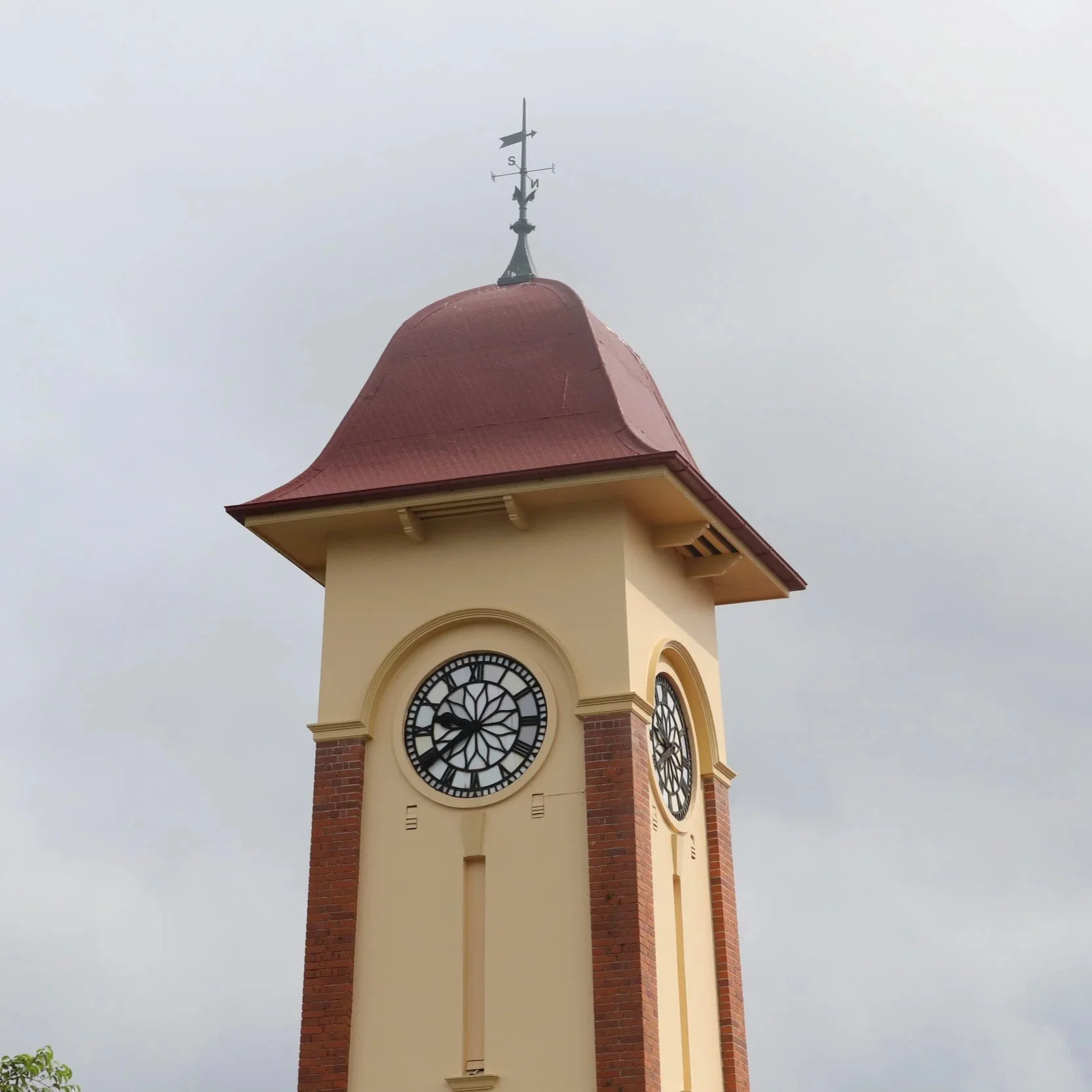 A clock tower with a red, curved roof and cream-colored walls, featuring brick accents, an ornate clock face, and a weather vane on top, against a cloudy sky.