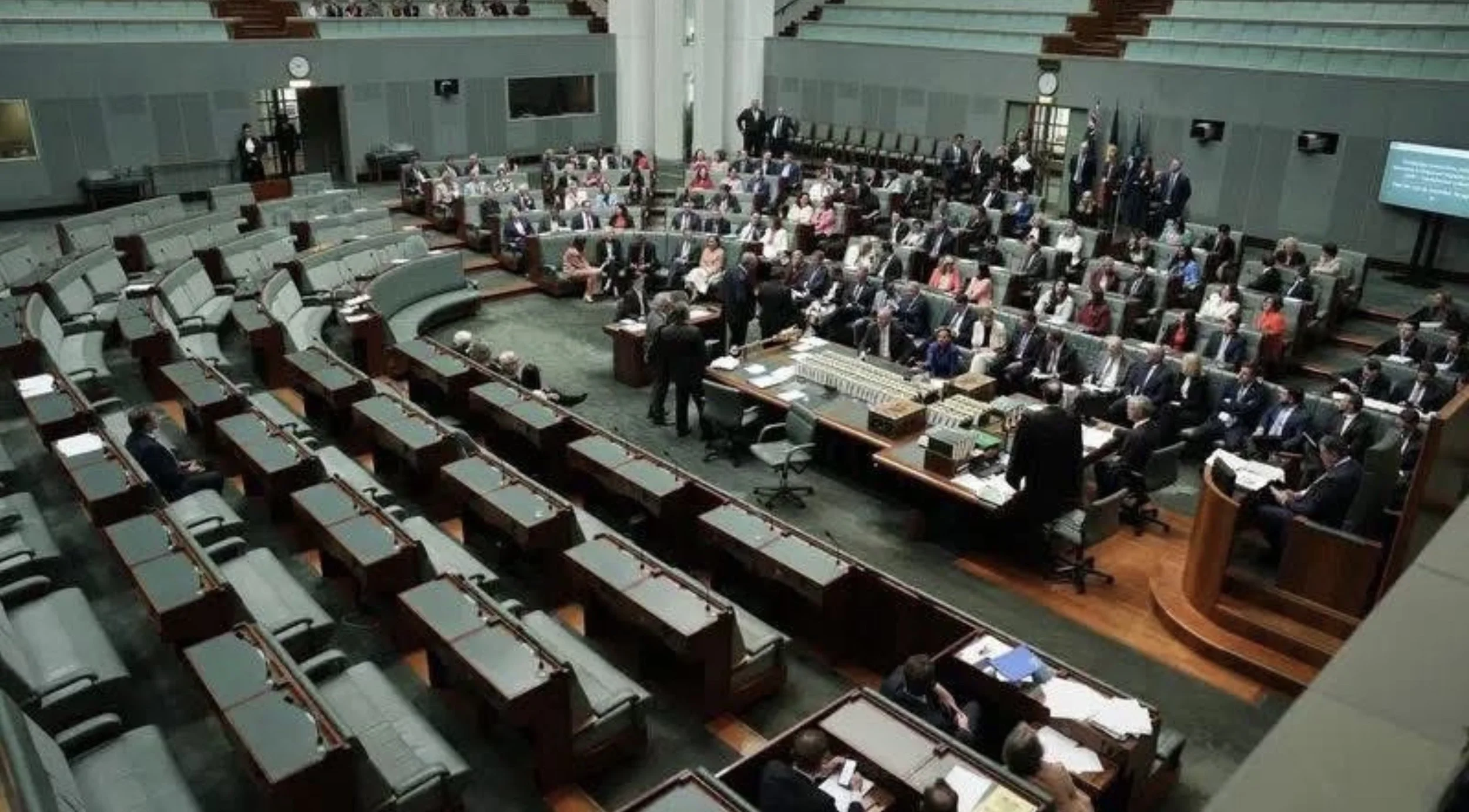 A legislative chamber with many seated people, possibly lawmakers, around a central speaking area with a raised platform, desks, and a few standing individuals.
