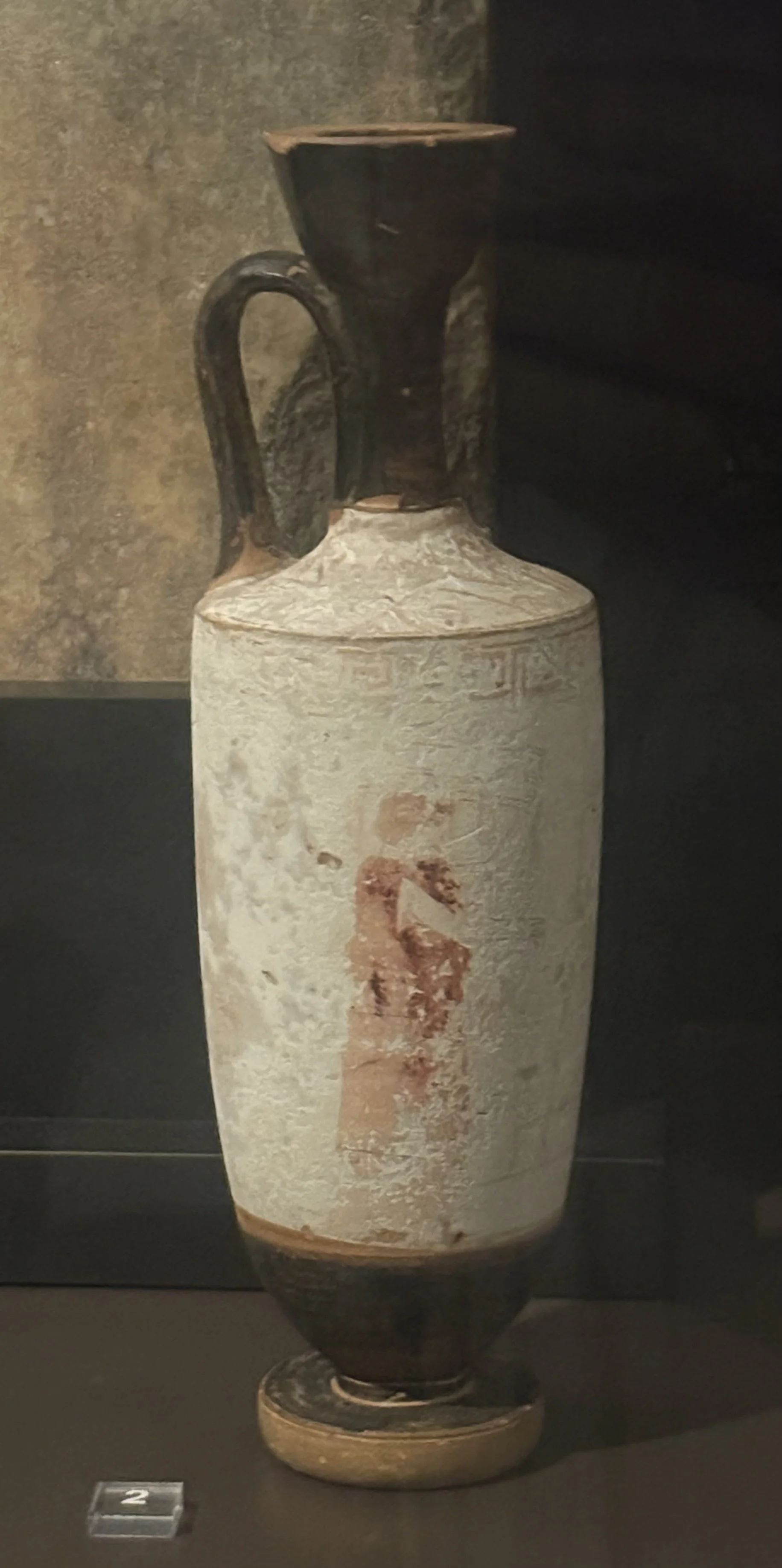 Ancient ceramic vase with a black top and handle, decorated with faded reddish-orange figures and patterns, displayed in a museum setting.