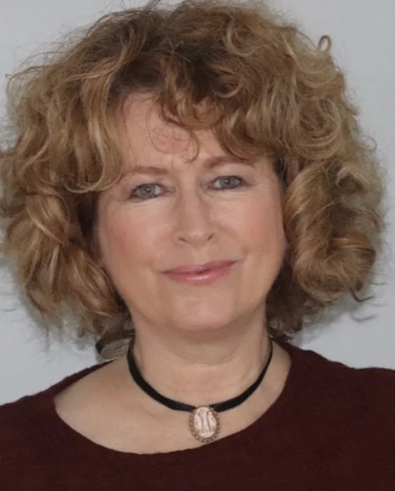 A woman with curly red hair smiling, wearing a dark top and a black choker necklace with a pendant.