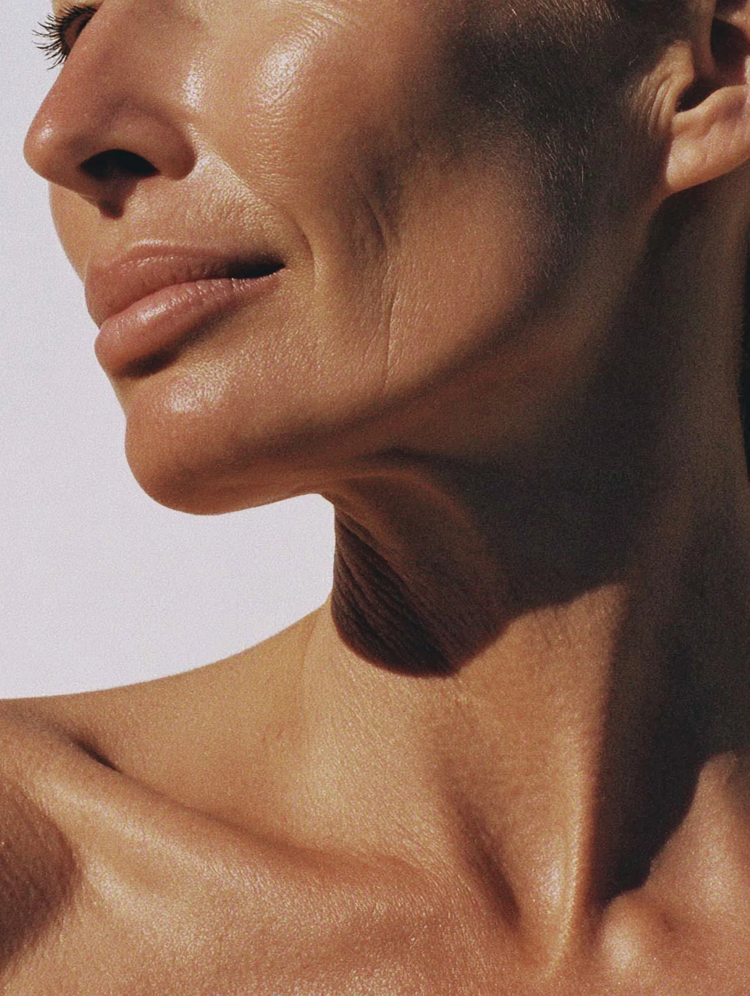 Close-up of a woman's face and shoulder, showing her smooth, tanned skin, lips, and part of her ear, with a neutral background.