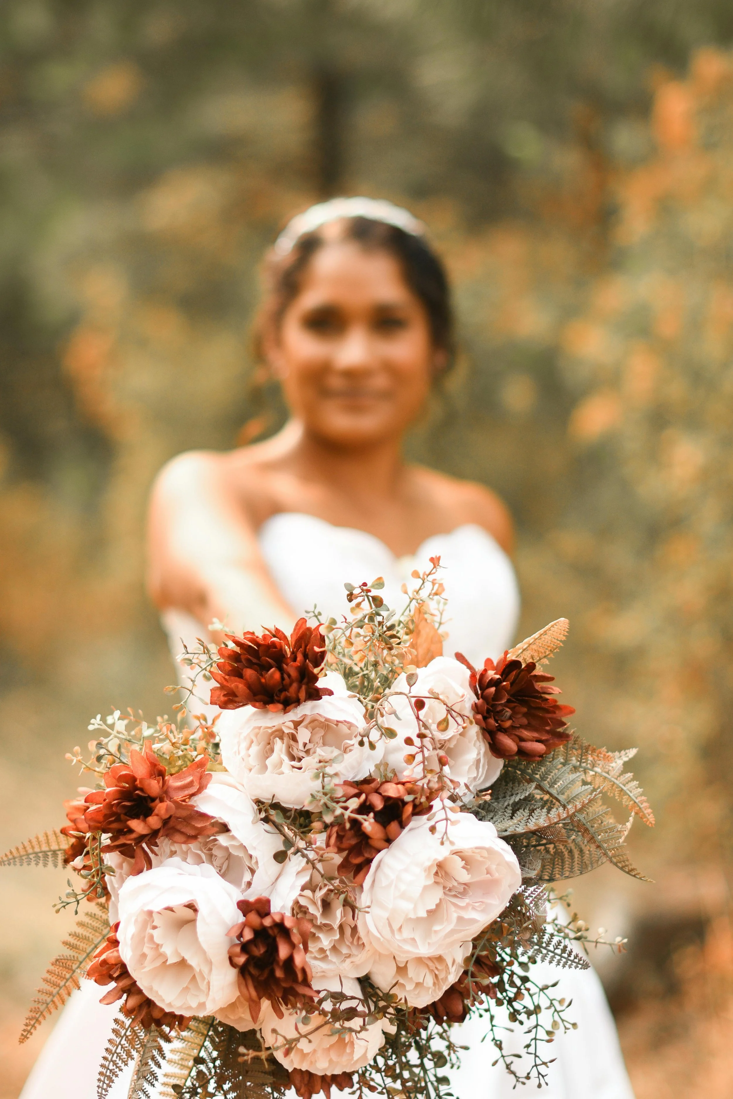 A woman in a white wedding dress holding a bouquet of white, peach, and brown flowers with greenery in an outdoor setting.