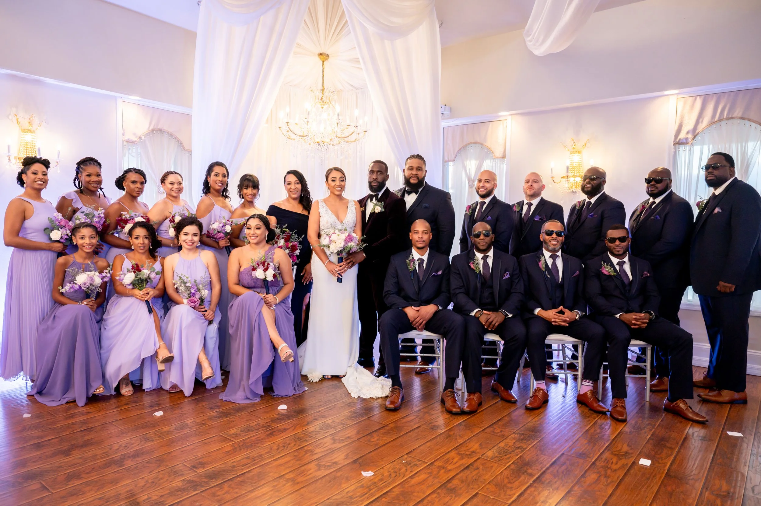 A wedding party with the bride and groom centered, surrounded by bridesmaids and groomsmen in formal attire, in an elegant room with draped white curtains, a chandelier, and hardwood floors.