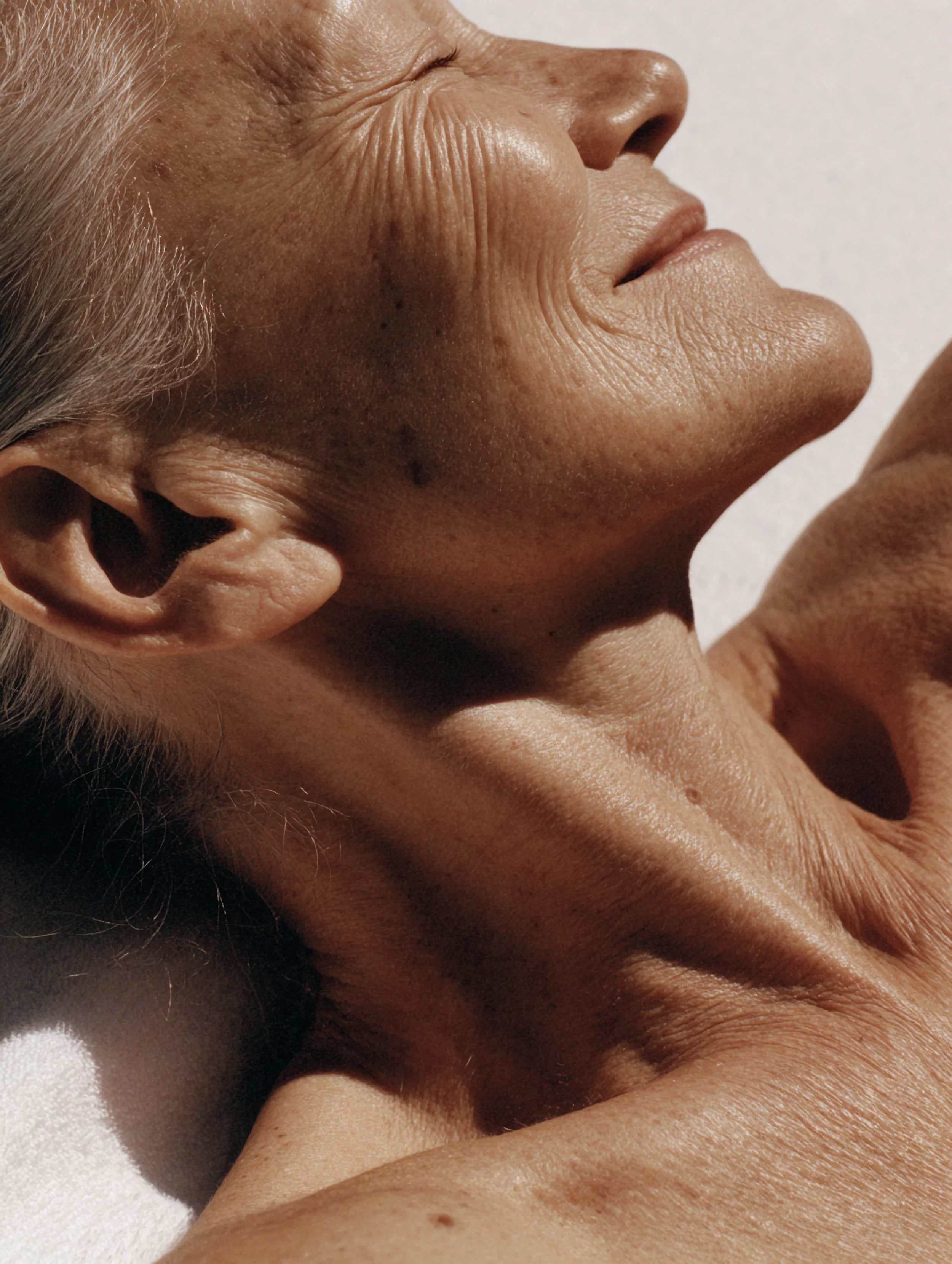Close-up of an elderly person's face and hand, showing skin with wrinkles and age spots, with part of their ear and lips visible.