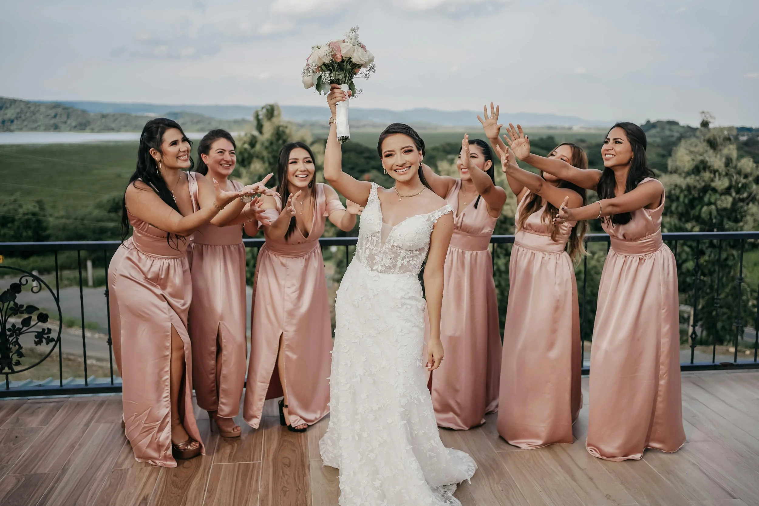 A bride in a white wedding dress celebrating with her bridesmaids, who are wearing matching blush pink dresses, on a balcony with scenic countryside in the background.