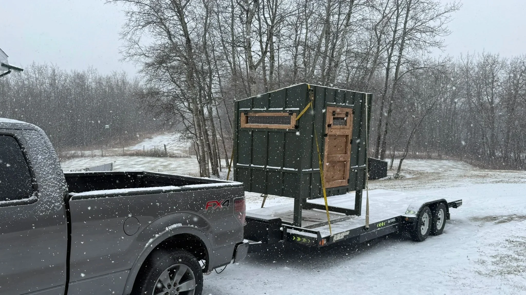 A black trailer attached to a gray pickup truck with snow on it. The trailer carries a small wooden shed with a window and door, set in a snowy outdoor landscape with bare trees and overcast sky.